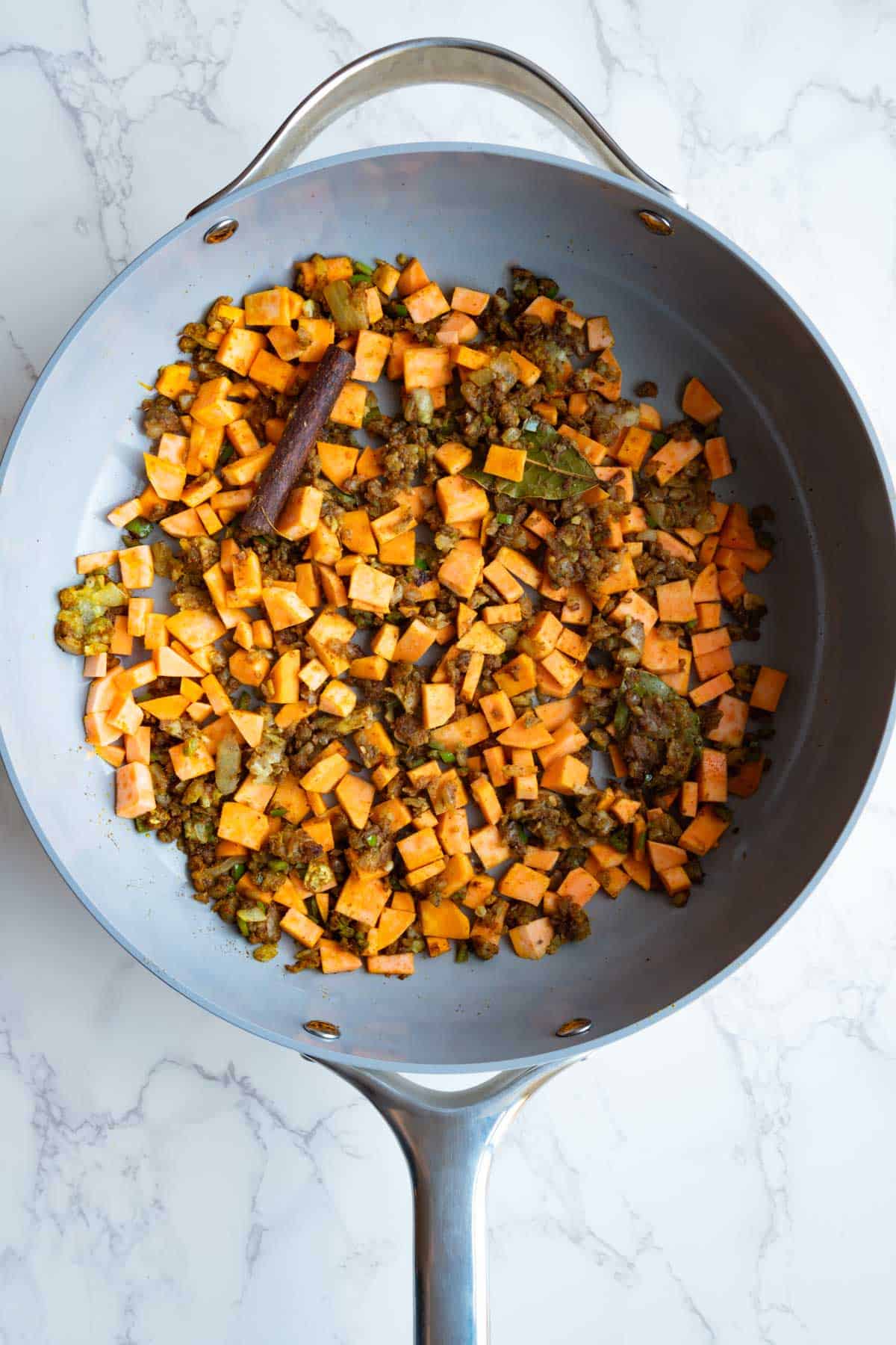 Cooked aromatics and seasoning, bay leaves, cinnamon stick, and cubed sweet potato in a large skillet.