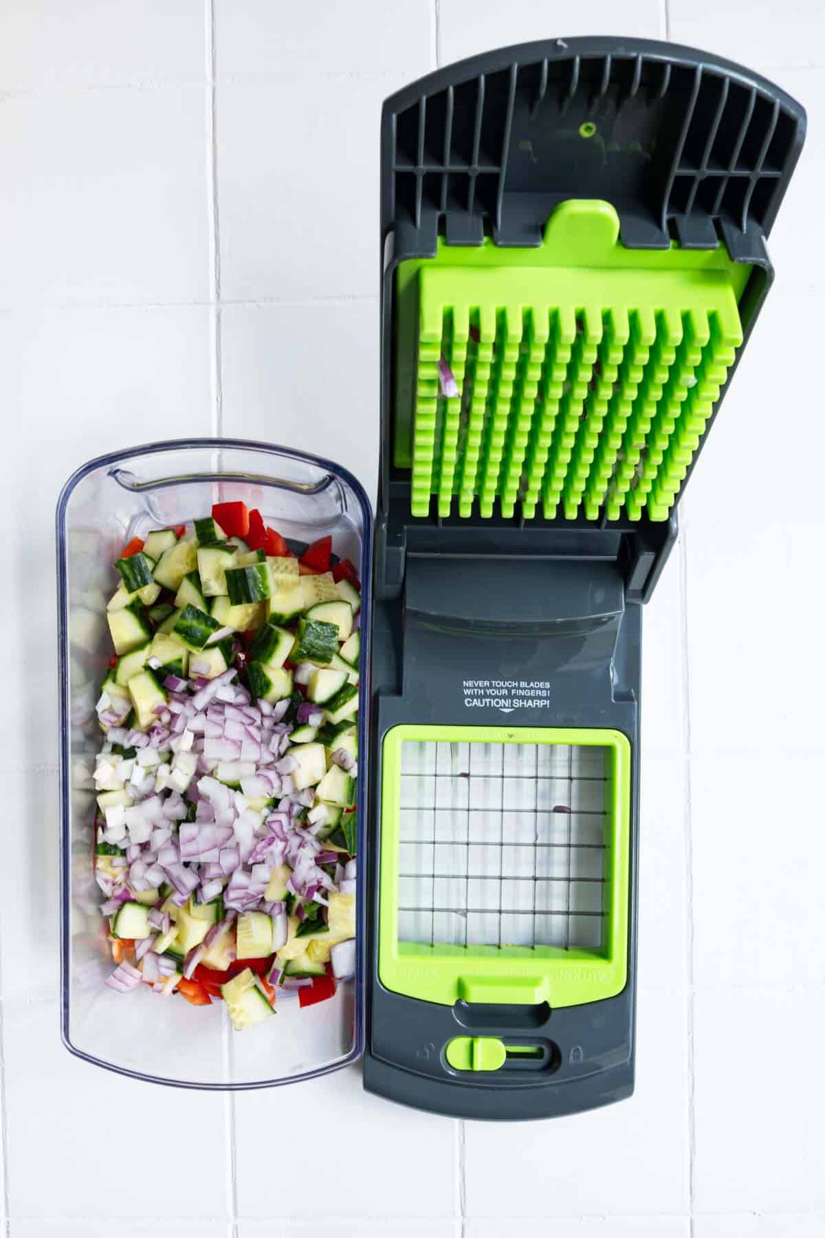 Chopped cucumber, red bell pepper, and red onion in a vegetable chopper.