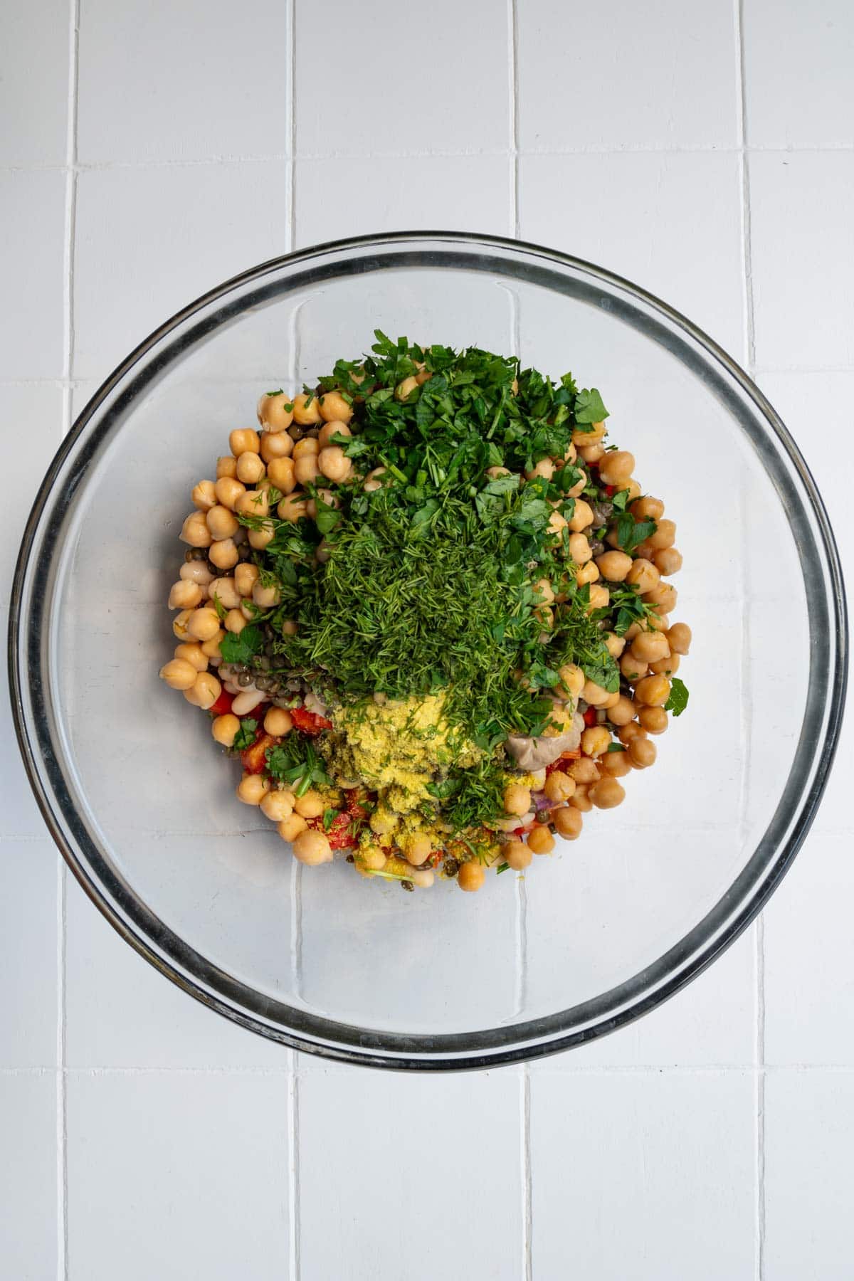 Chickpeas, lentils, and white beans with red bell pepper, fresh herbs, nutritional yeast, and mustard in a large glass bowl.