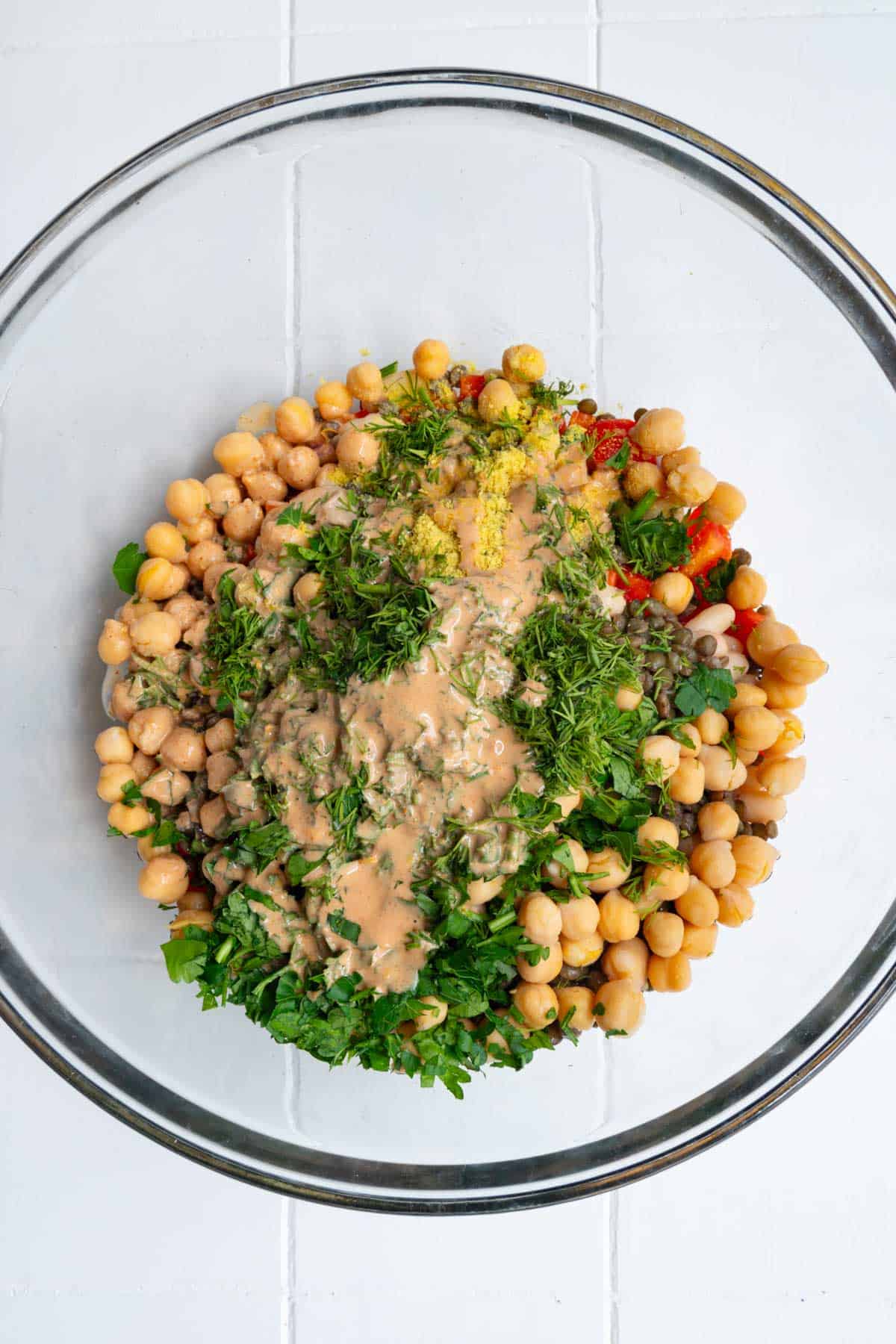 Chickpeas, lentils, and white beans with red bell pepper, fresh herbs, tahini, lemon juice, and seasonings in a large glass bowl.