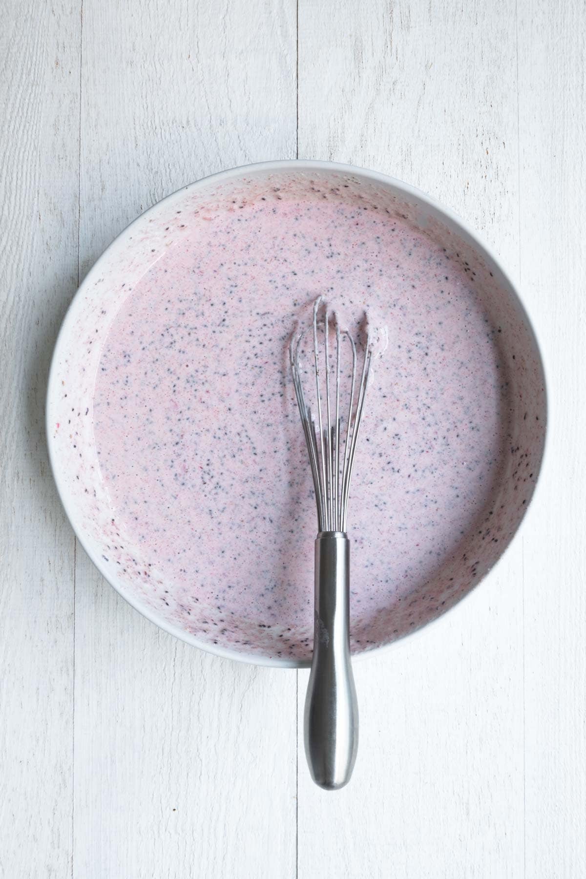 Ground and whole chia seeds with nondairy milk, yogurt, cooked and mashed strawberries and dates in a white bowl with a whisk.