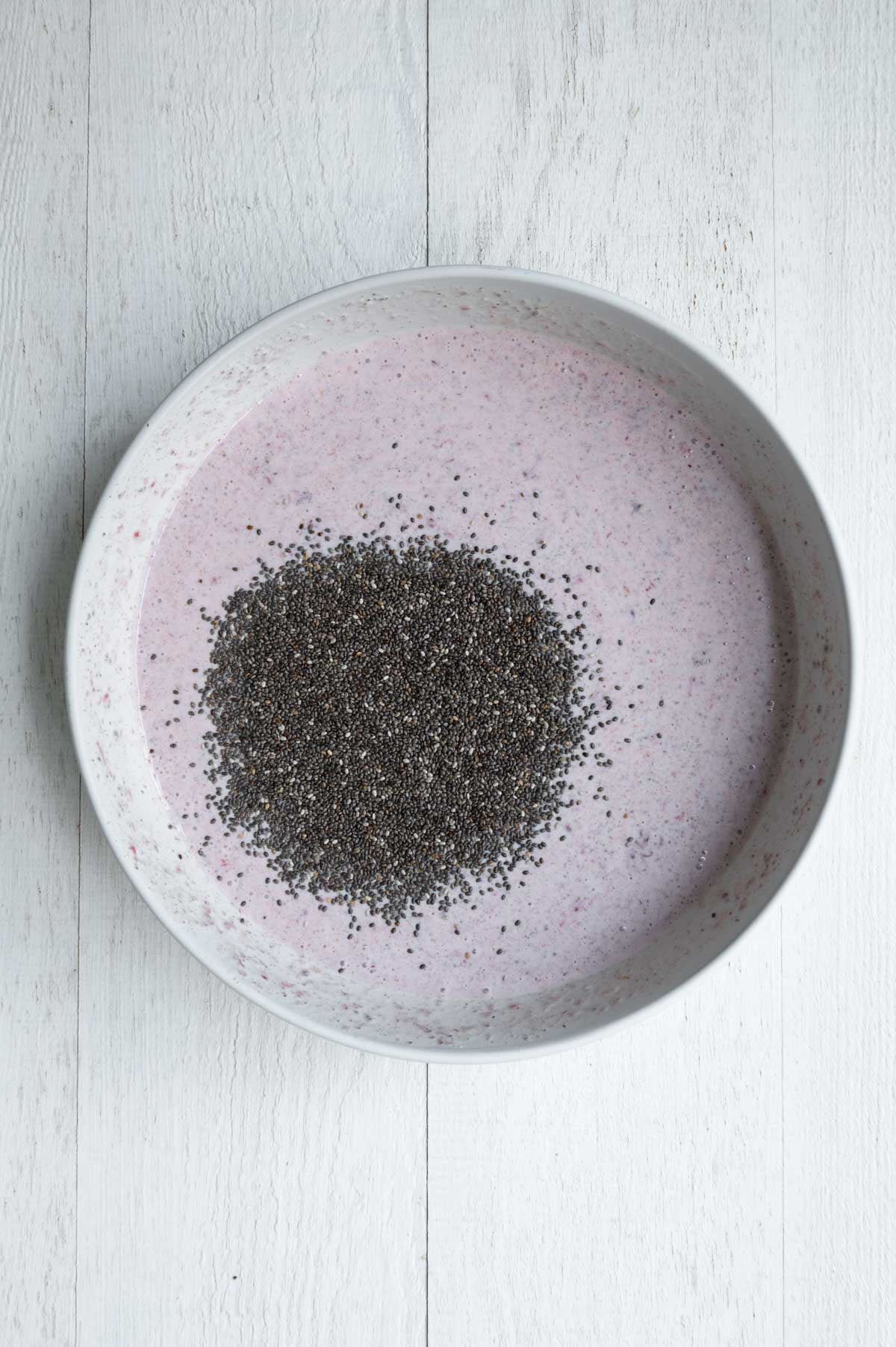 Ground chia seeds mixed into nondairy milk, yogurt, cooked and mashed strawberries and dates in a white bowl topped with whole chia seeds.