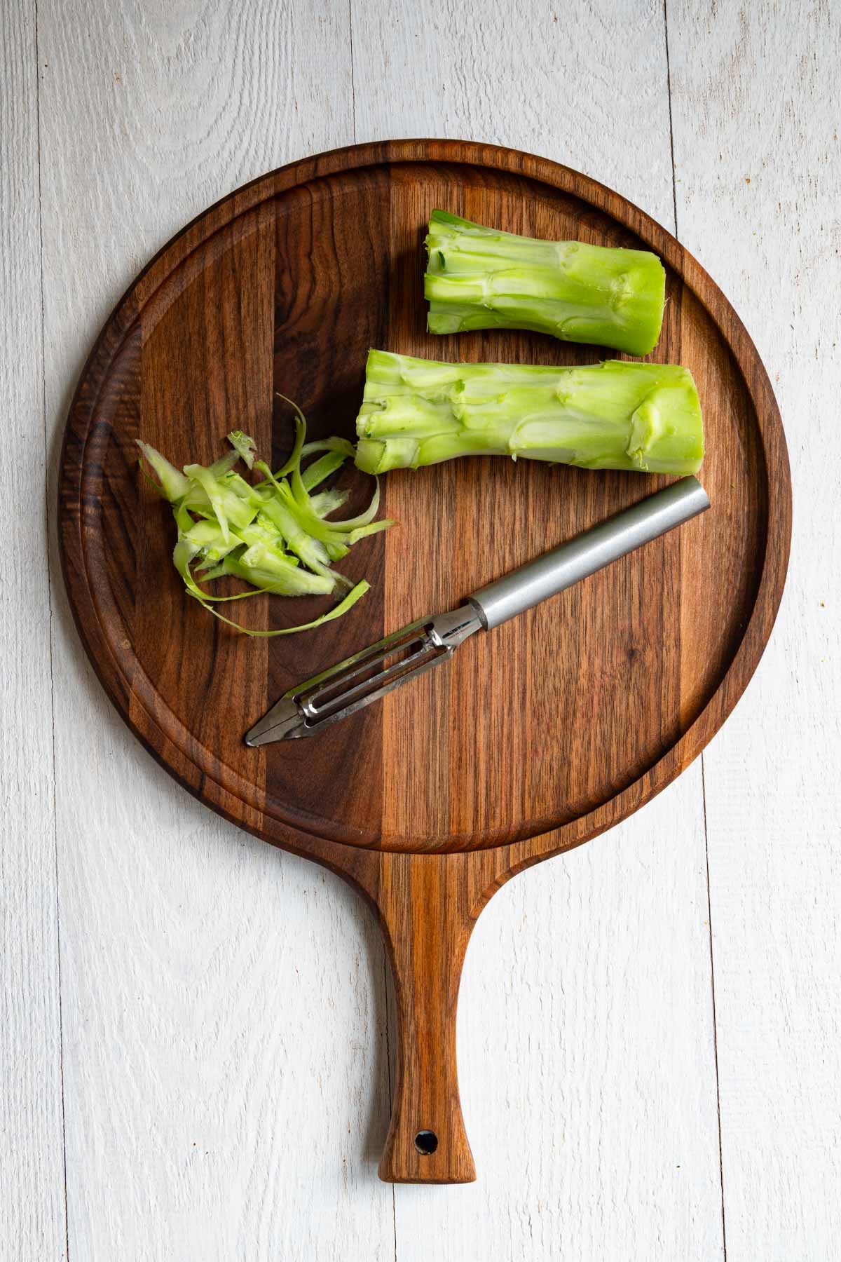 Peeled broccoli stems in a wooden cutting board with a peeler.