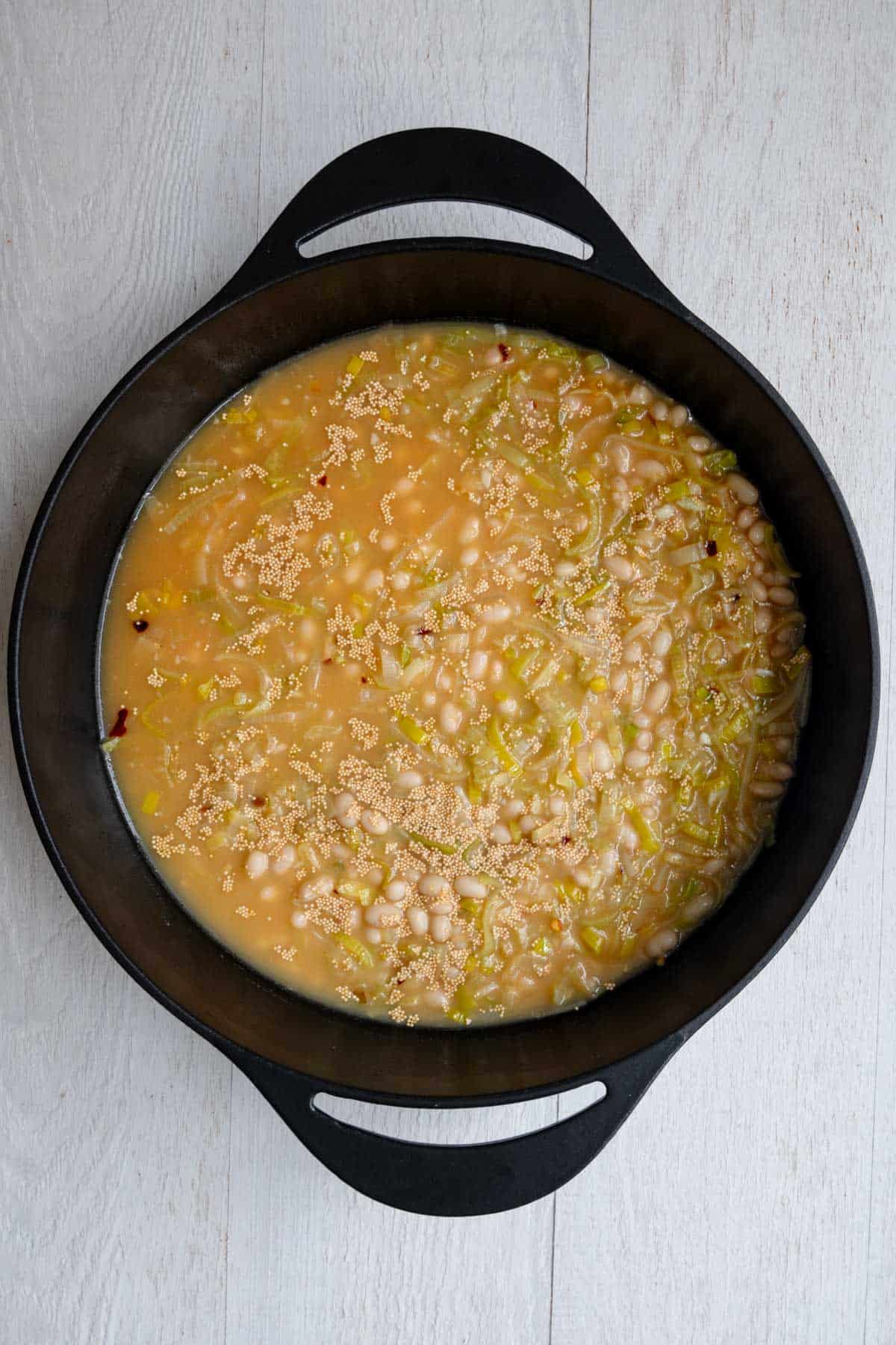 Vegetable broth with beans, amaranth, chopped leek, crushed red pepper, and garlic cooking in a cast-iron skillet.