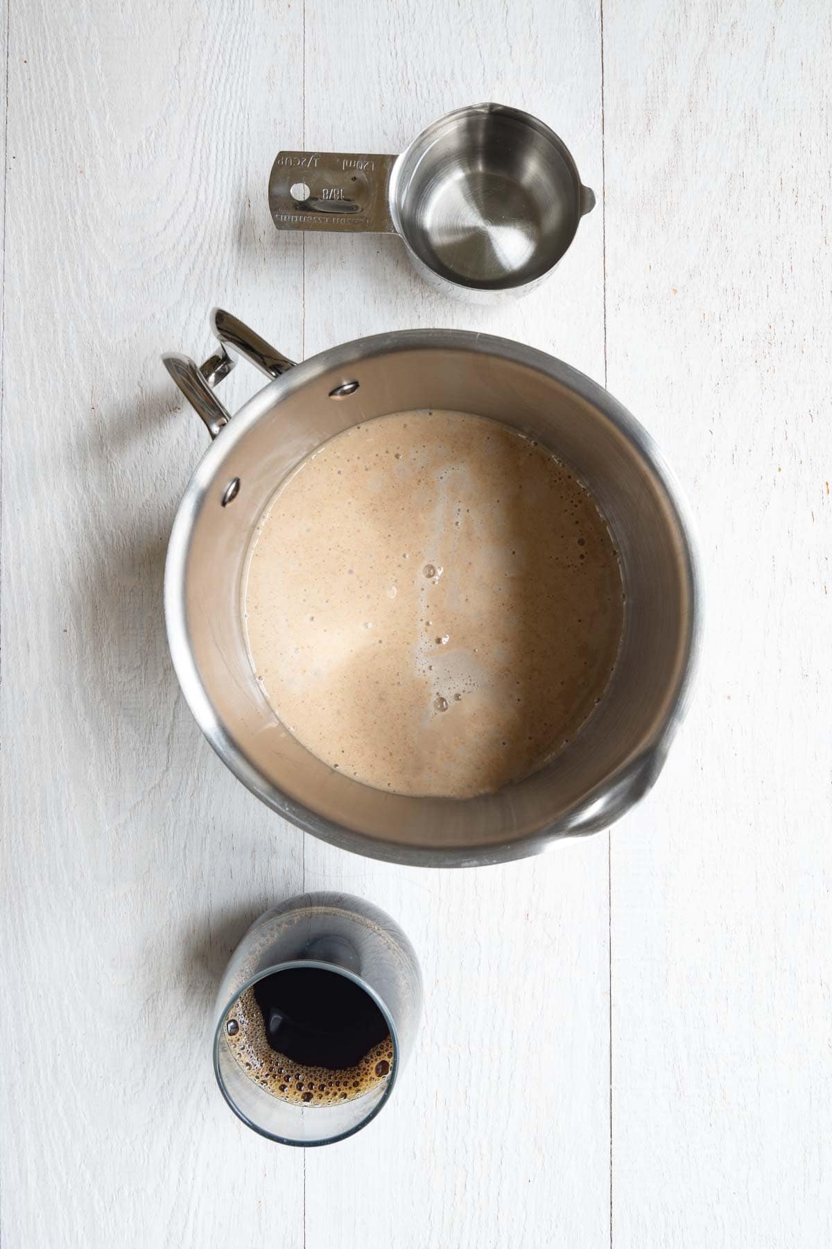 Heated latte in a stainless steel saucepan with a pouring spout and a cup of coffee and measuring cup of water.