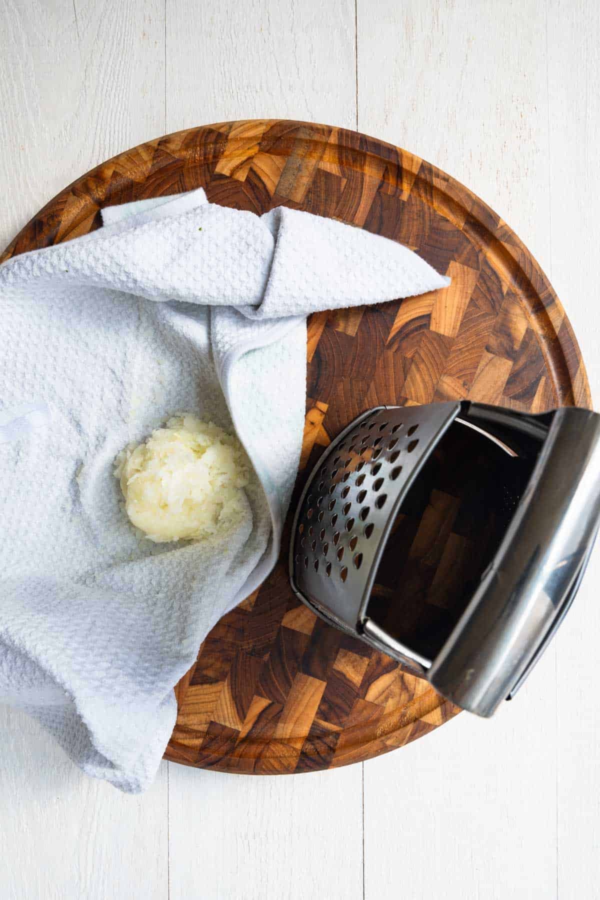 Grated onion in a kitchen towel next to a grater on a wooden cutting board.