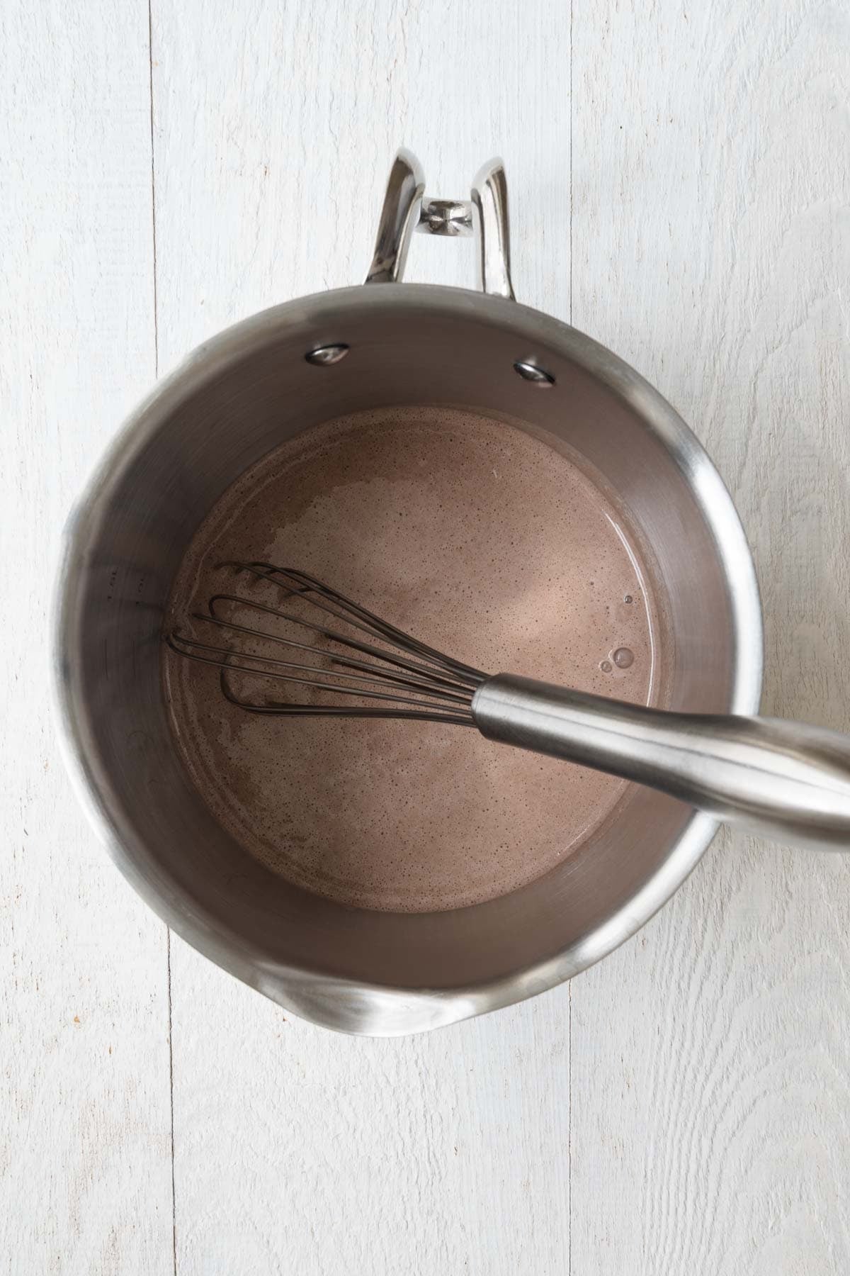 Brown creamy liquid in a small saucepan with a spout and a whisk.