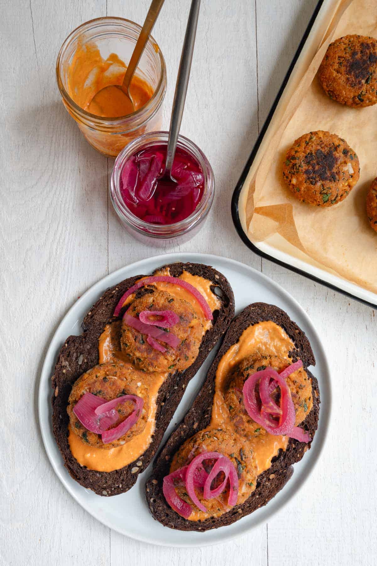 Gluten-free tofu burgers served on pumpernickel toast with orange sauce and pickled red onion and leftovers on a baking sheet.