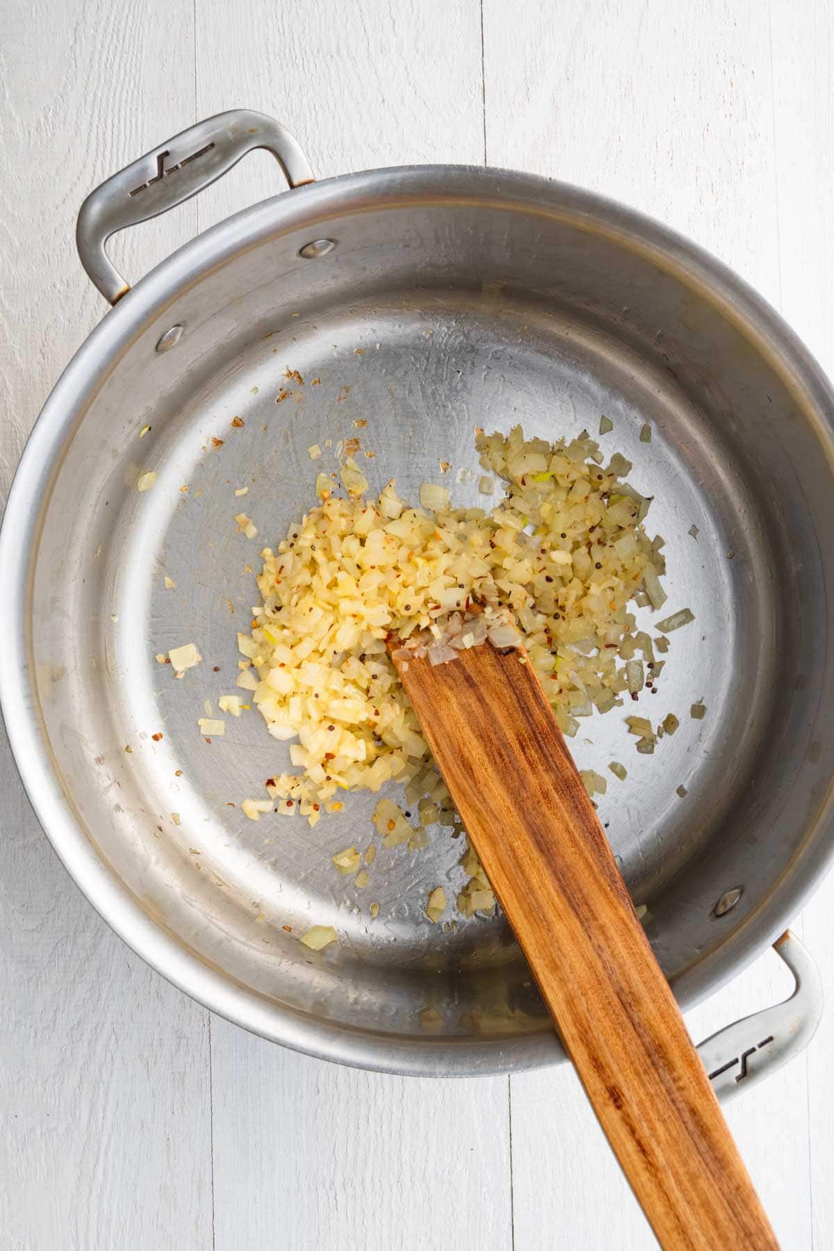 Chopped onion and garlic cooking with crushed red pepper and seasoning in a stainless steel saucepan.