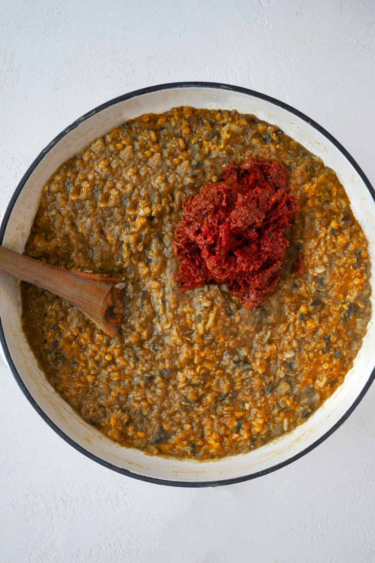 Eggplant, red lentils, broth, onion, garlic, and red sauce in a large white skillet with a wooden spoon.
