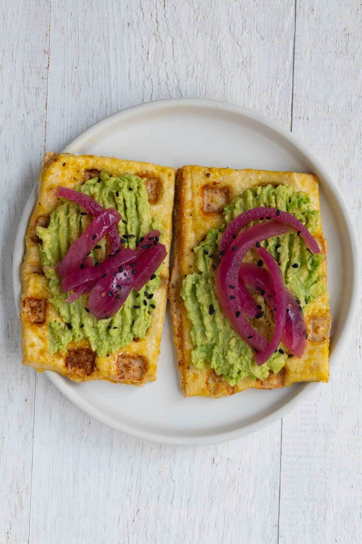Tofu waffles on a white plate topped with avocado, pickled red onion, and sesame seeds.