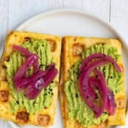 Tofu waffles on a plate topped with avocado and pickled red onion.