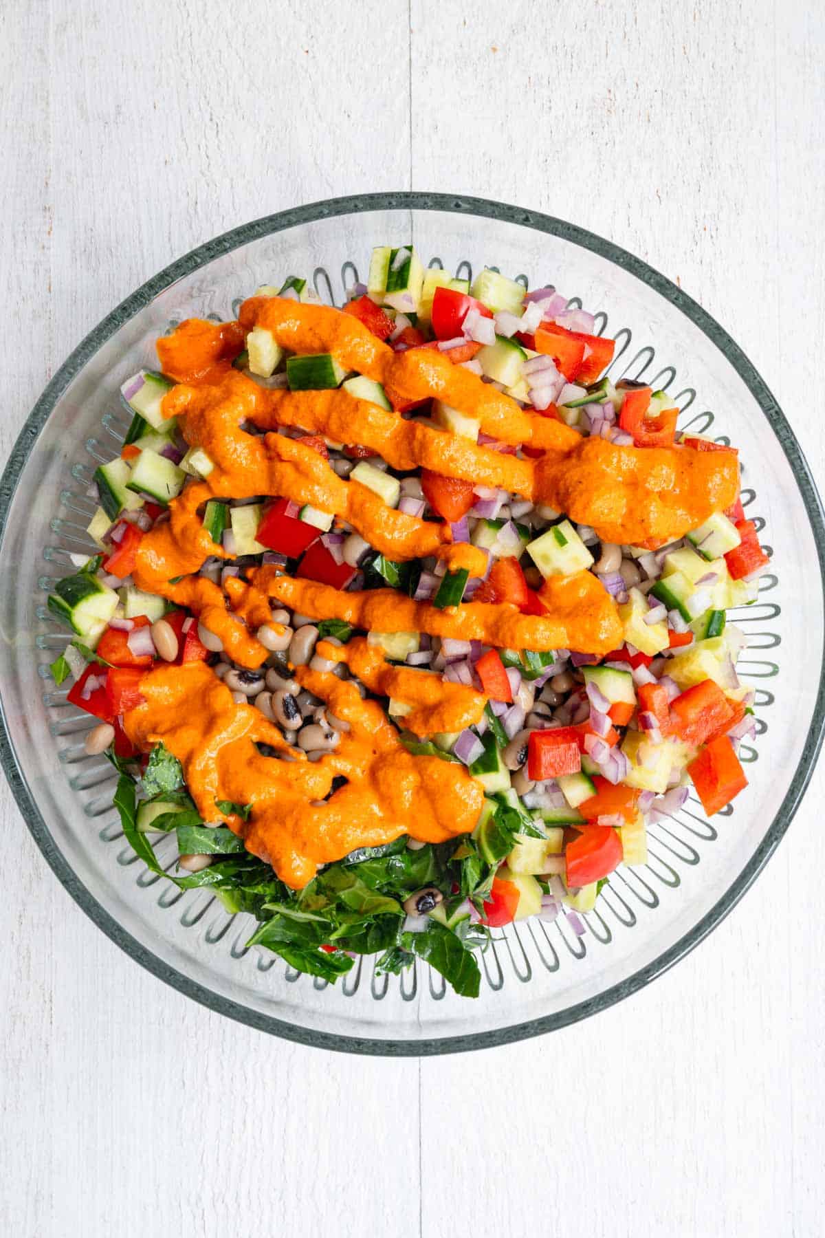 Chopped cucumber, red bell pepper, red onion, collard greens, and black eyed peas in a glass bowl topped with roasted bell pepper sauce.