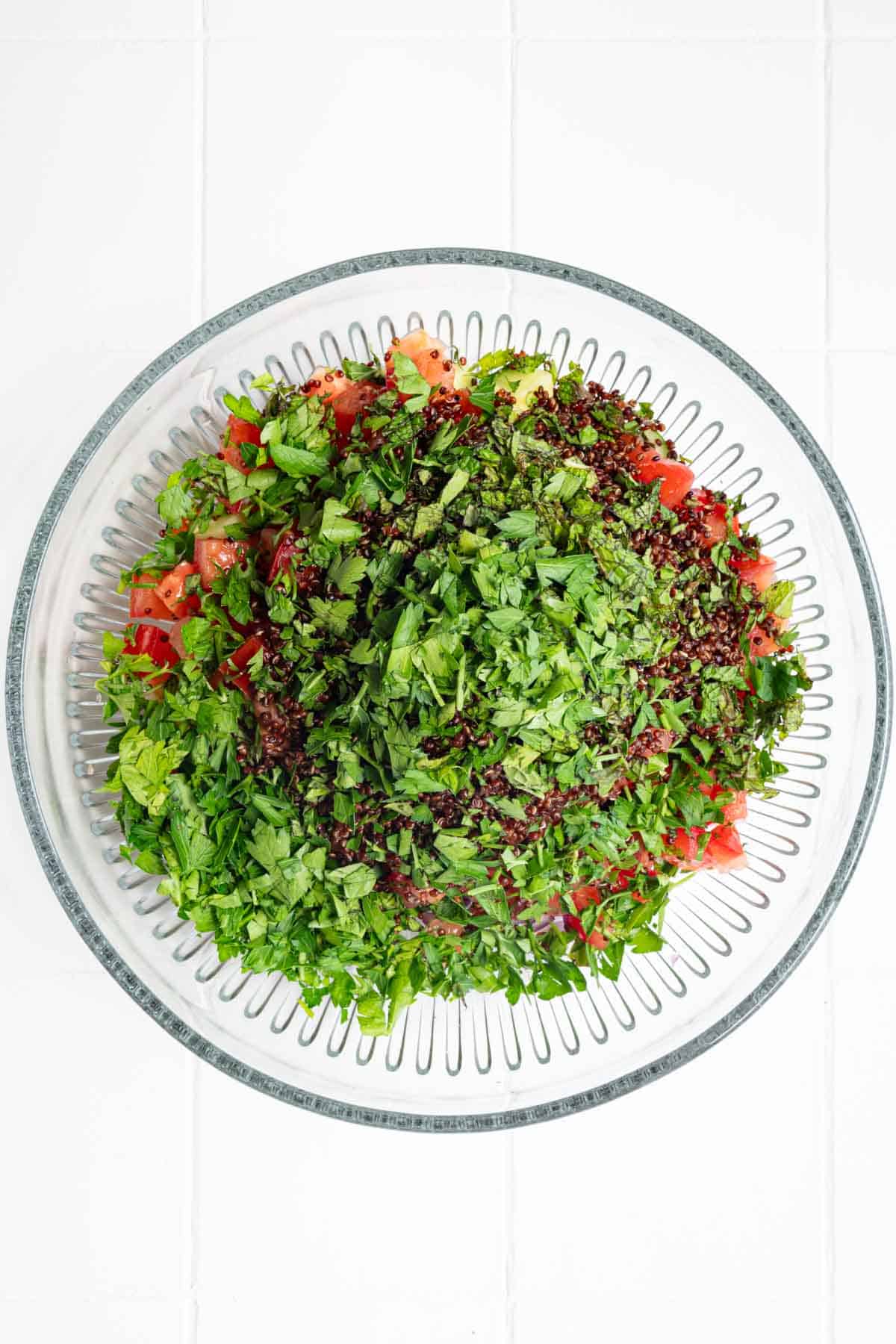 Red quinoa, chopped tomatoes, cucumbers, red onion, parsley, and mint in a glass bowl.