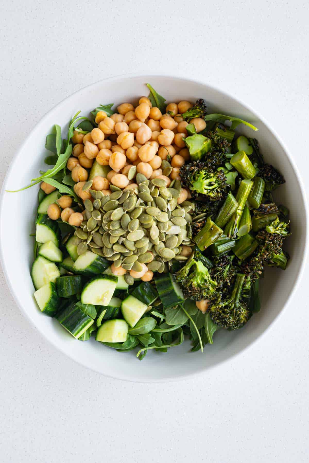 Arugula, chickpeas, roasted broccolini, cucumber, and pumpkin seeds in a bowl.