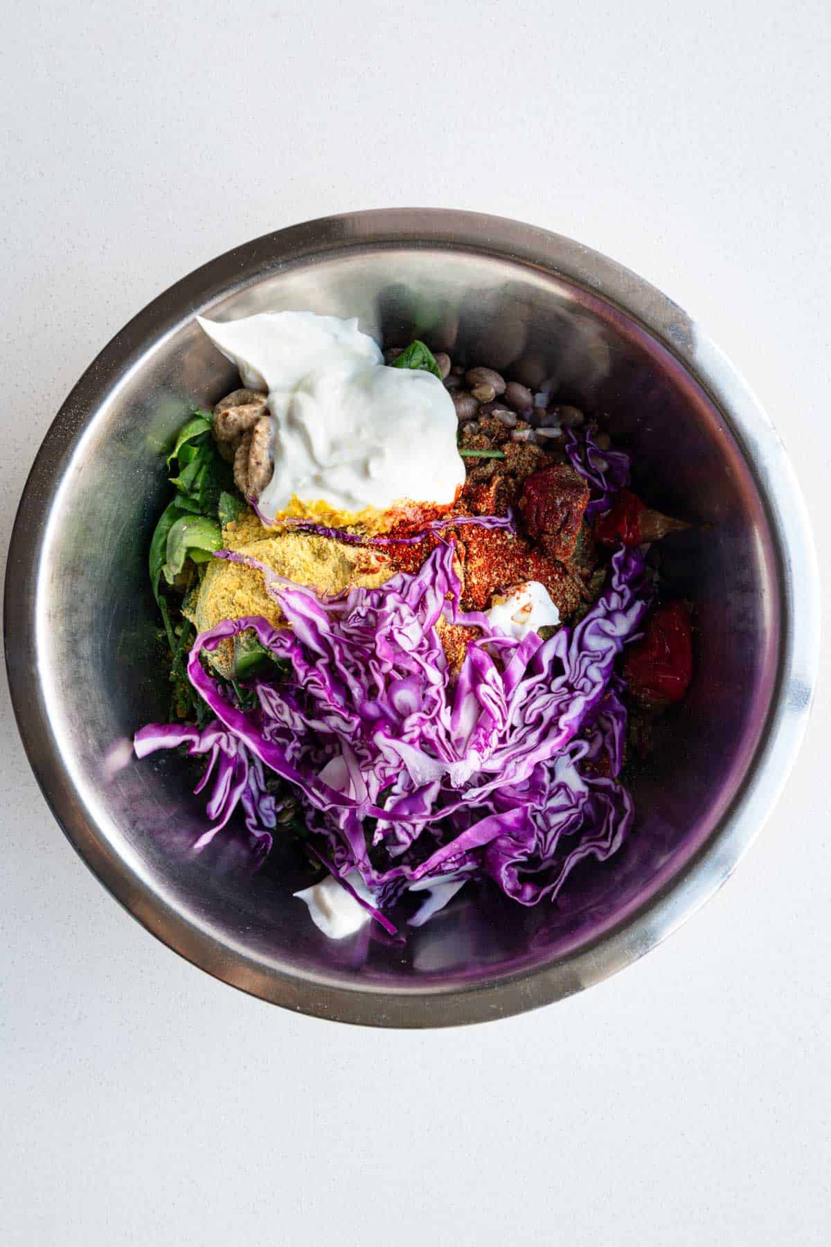 Lentils, beans, red cabbage, yogurt, lemon, Calabrian chilis, herbs, red onion, mustard, capers, red onion, and nutritional yeast in a metal bowl.