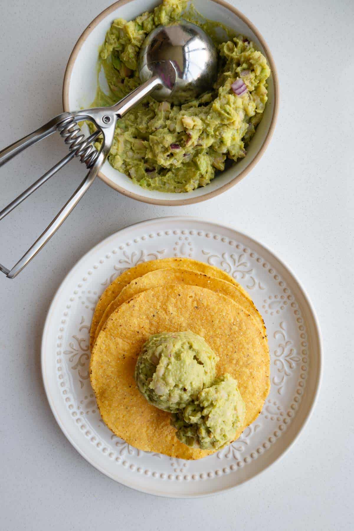 Green mash in a bowl with an ice cream scoop and on a baked corn tortilla topped with sprouts.