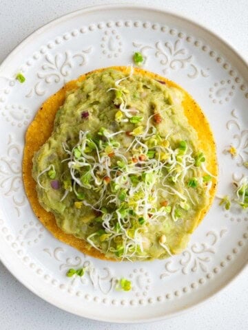 White bean mash with avocado on a tostada topped with sprouts.