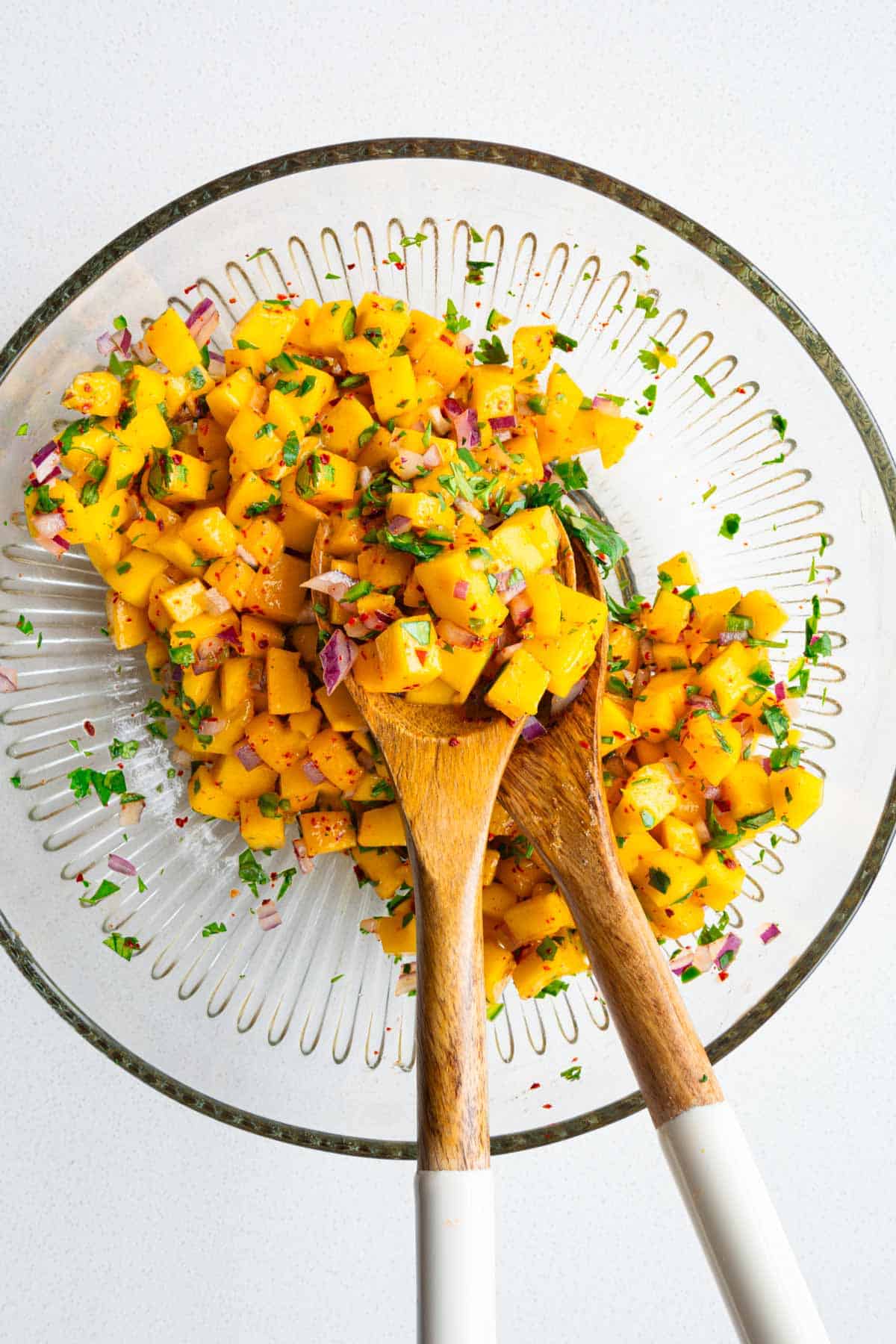 Chopped mango, red onion, jalapeño, lime juice, red pepper powder, and cilantro in a bowl mixed with salad tongs.