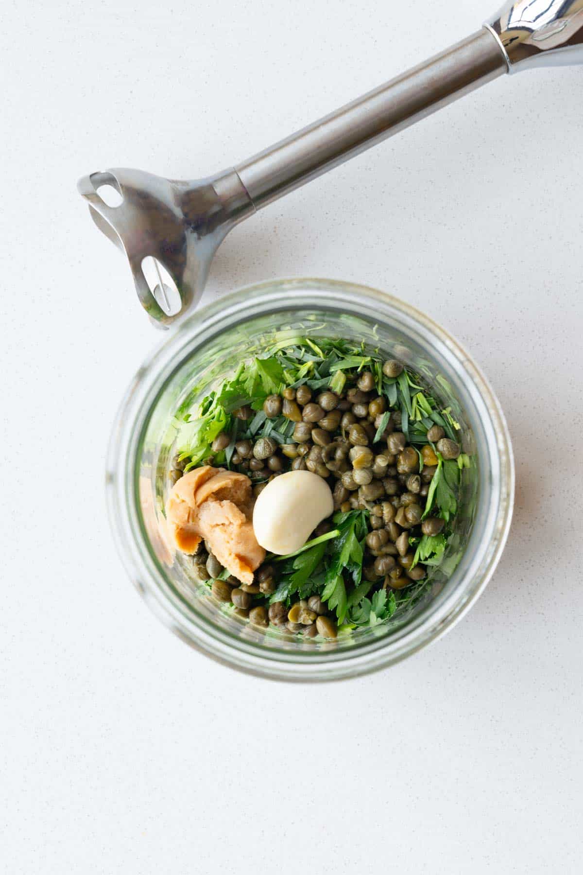 Capers, pumpkin seeds, tarragon, chives, parsley, miso, garlic, tahini, and lemon in a glass jar with immersion blender.