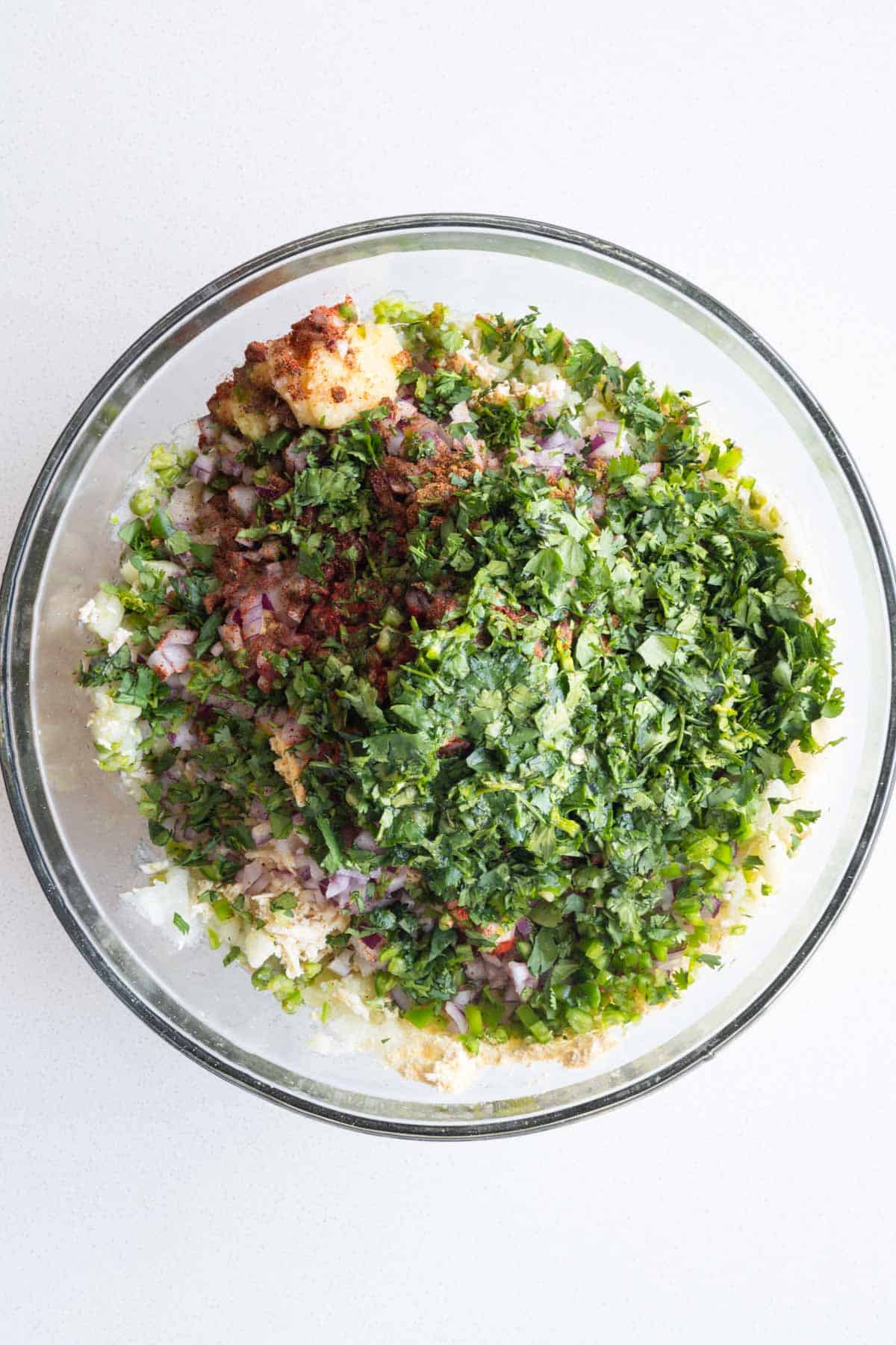 Grated tofu, mashed potatoes, green chilis, ginger, garlic, mashed green peas, lemon juice, seasonings, chickpea flour, and cilantro in a glass bowl.