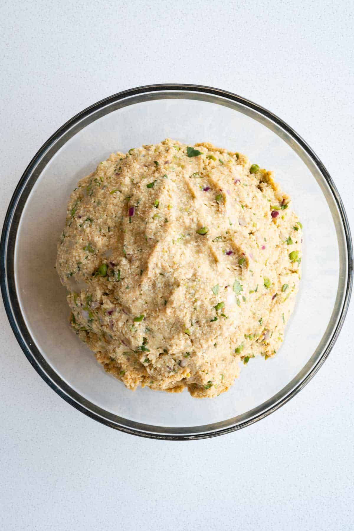 Tofu, potato, seasoning, chickpea flour, aromatics, and fresh herbs pressed intoa ball in a glass bowl.