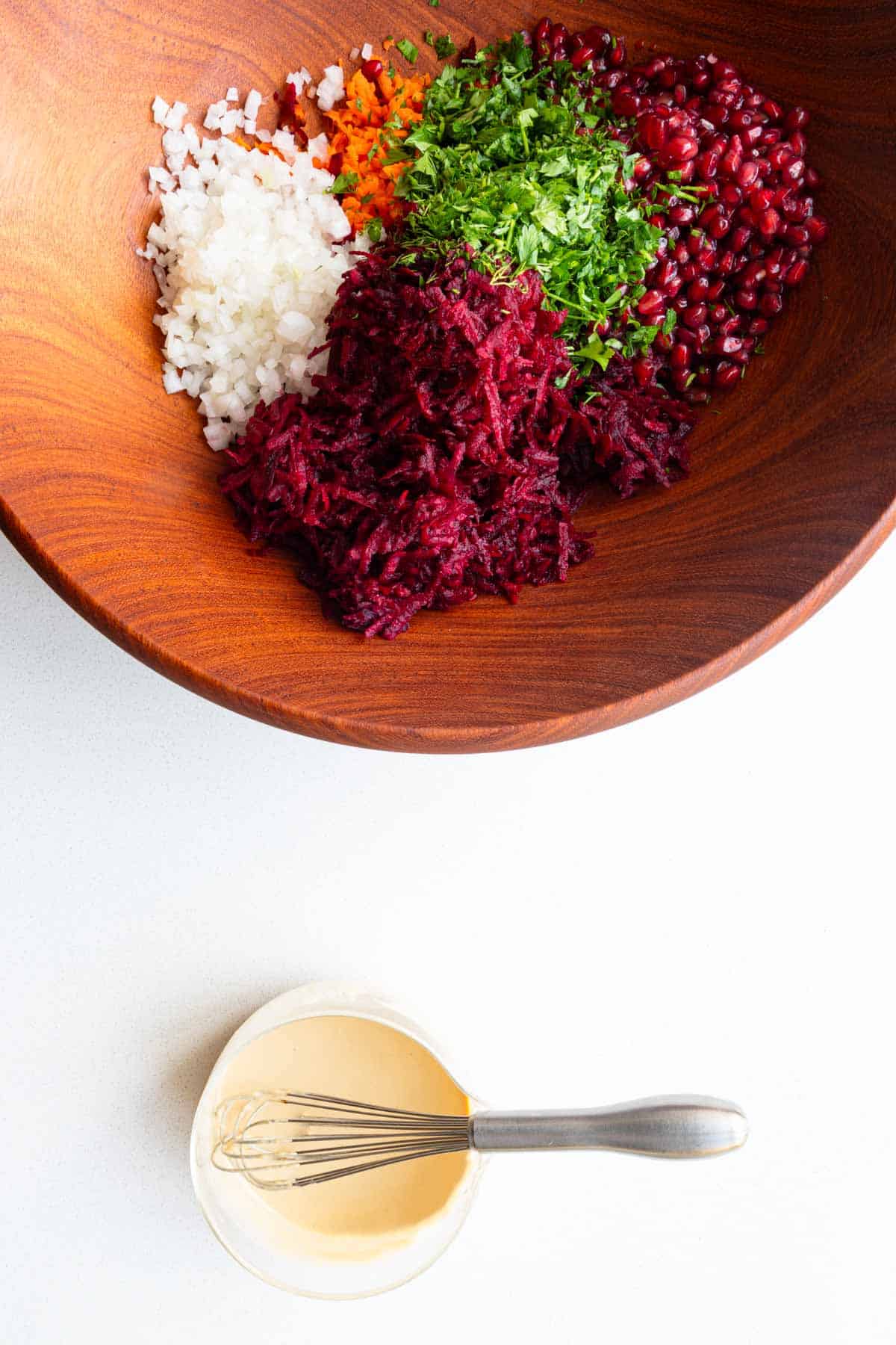 Grated beets and carrots with pomegranate arils, fresh herbs, and chopped onion in a wooden bowl next to whisked tahini dressing.