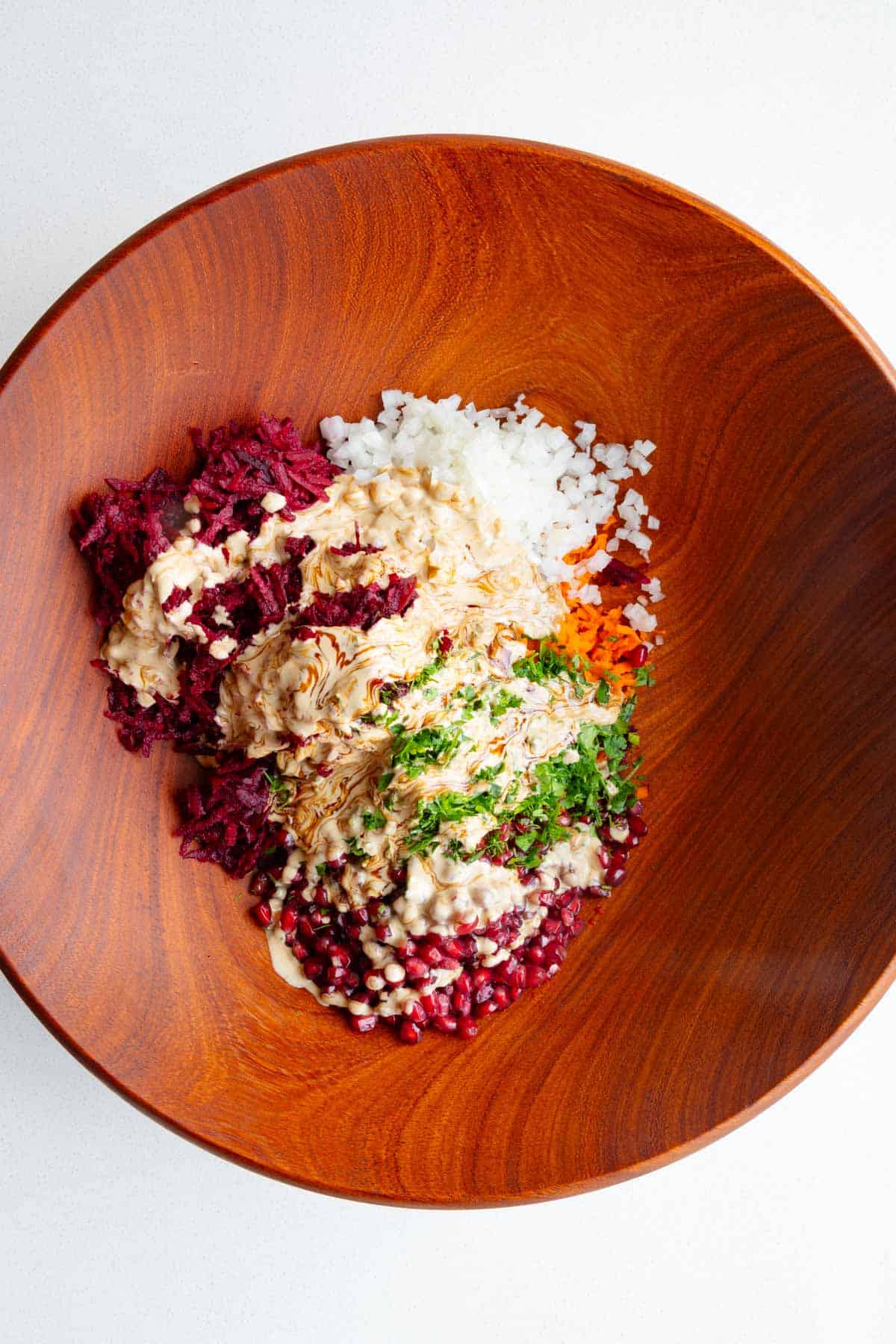 Beetroot Salad with Pomegranate, carrots, fresh herbs, onion, pumpkin seeds, and tahini dressing in a wooden bowl.