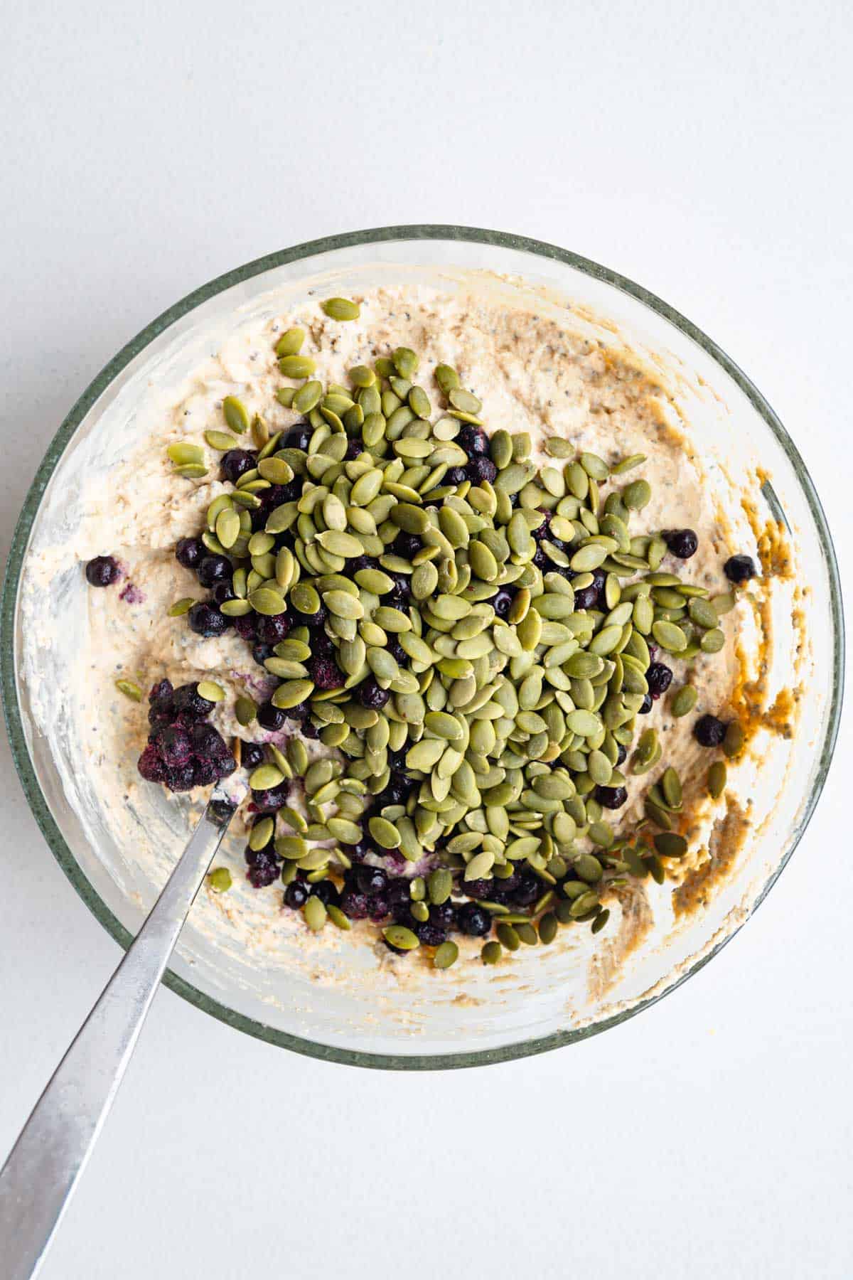 Mixed muffin batter topped with pumpkin seeds and blueberries in a glass bowl.