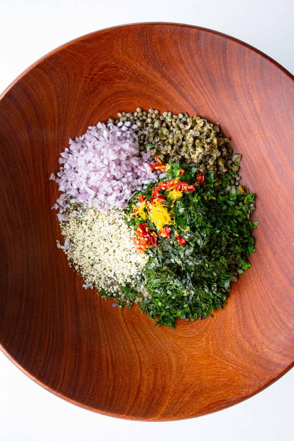 Lentils, chopped bell pepper, fresh herbs, shallots, capers, lemon zest, hemp hearts, Calabrian chilis, and red wine vinegar in a wooden bowl.