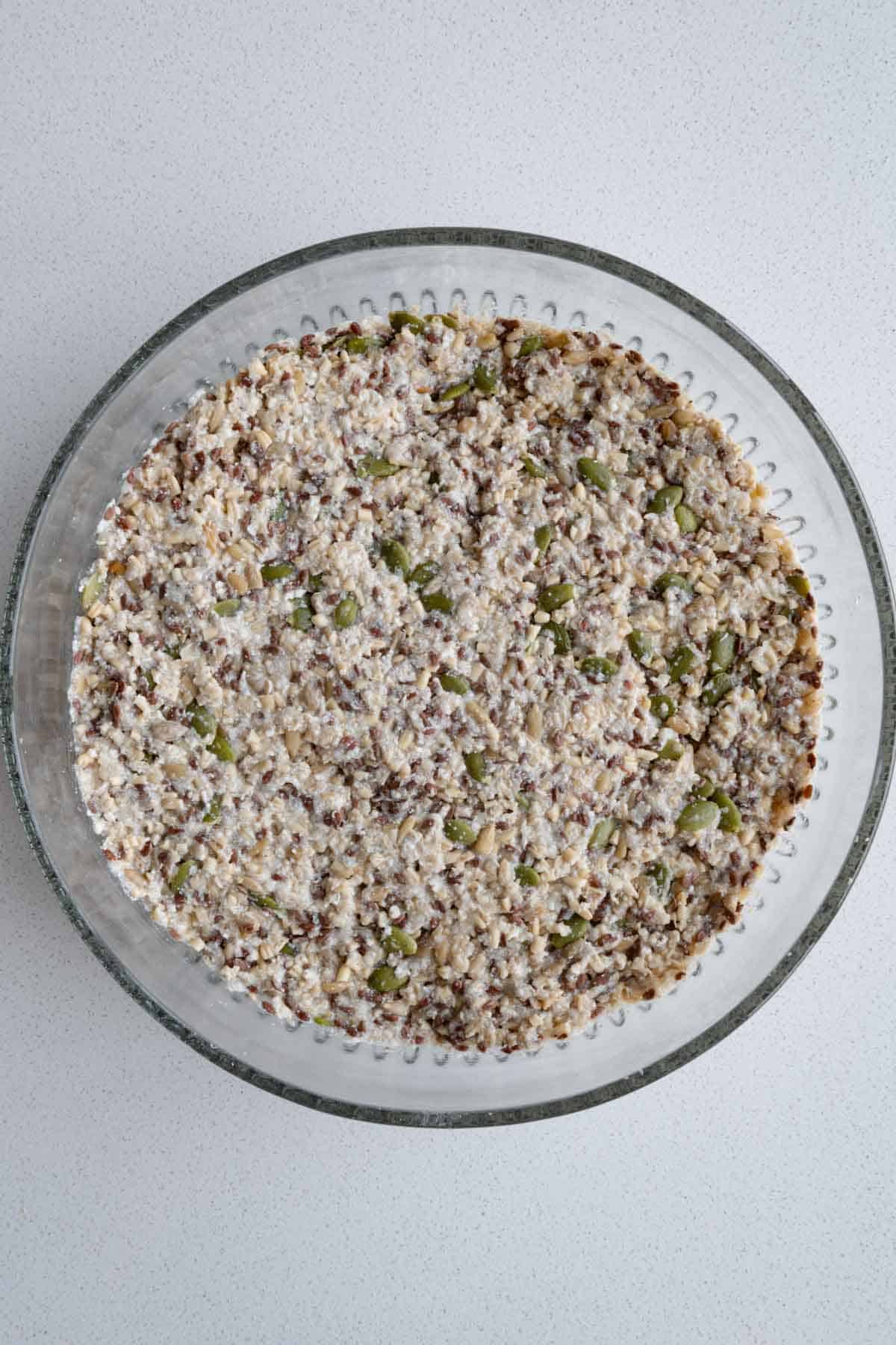 Drained soaked steel cut oats, pumpkin seeds, sunflower seeds, flax seeds, quinoa flakes, psyllium husks, water, and apple cider vinegar mixed into a dough in a glass bowl.