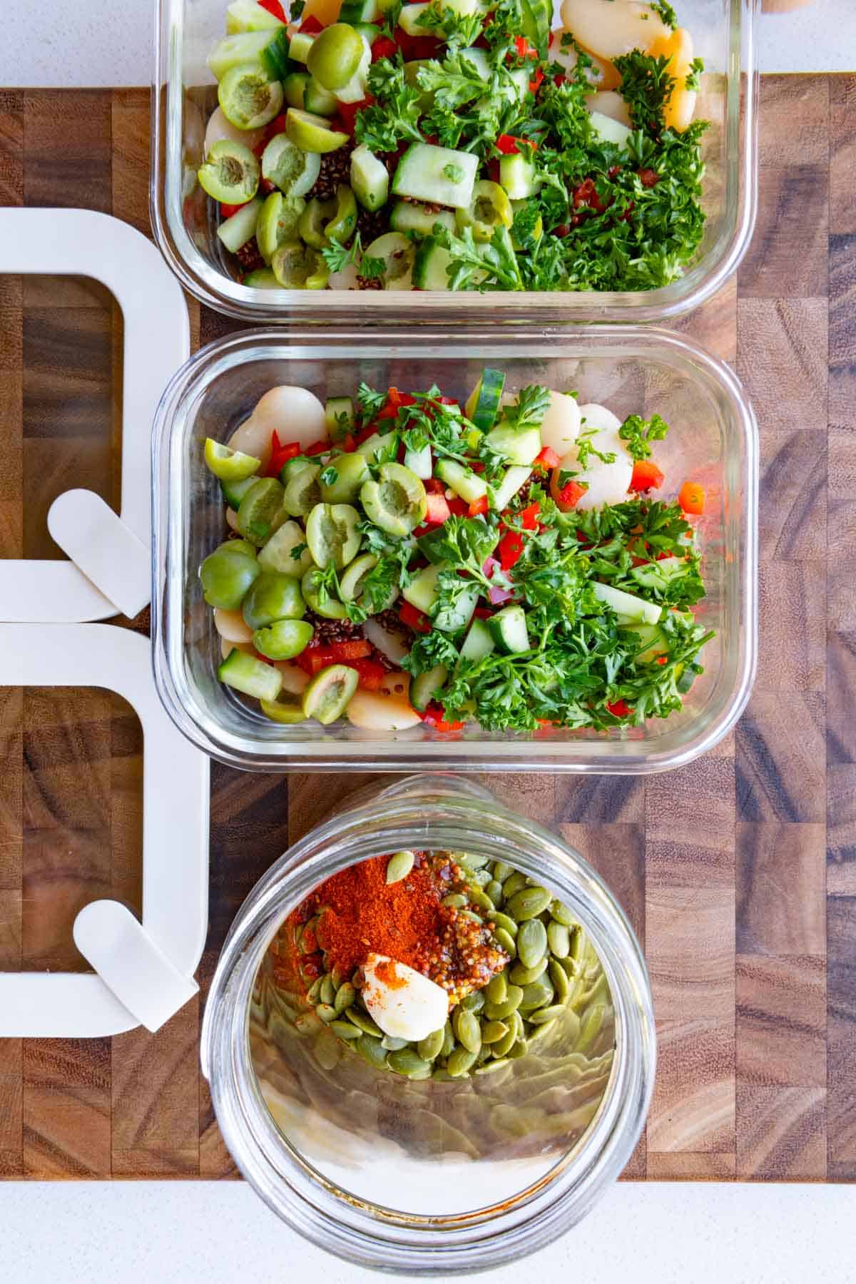 Butter beans, red quinoa, chopped herbs, scallions, and bell pepper in a two meal prep containers, and pumpkin seed dressing in a glass jar.