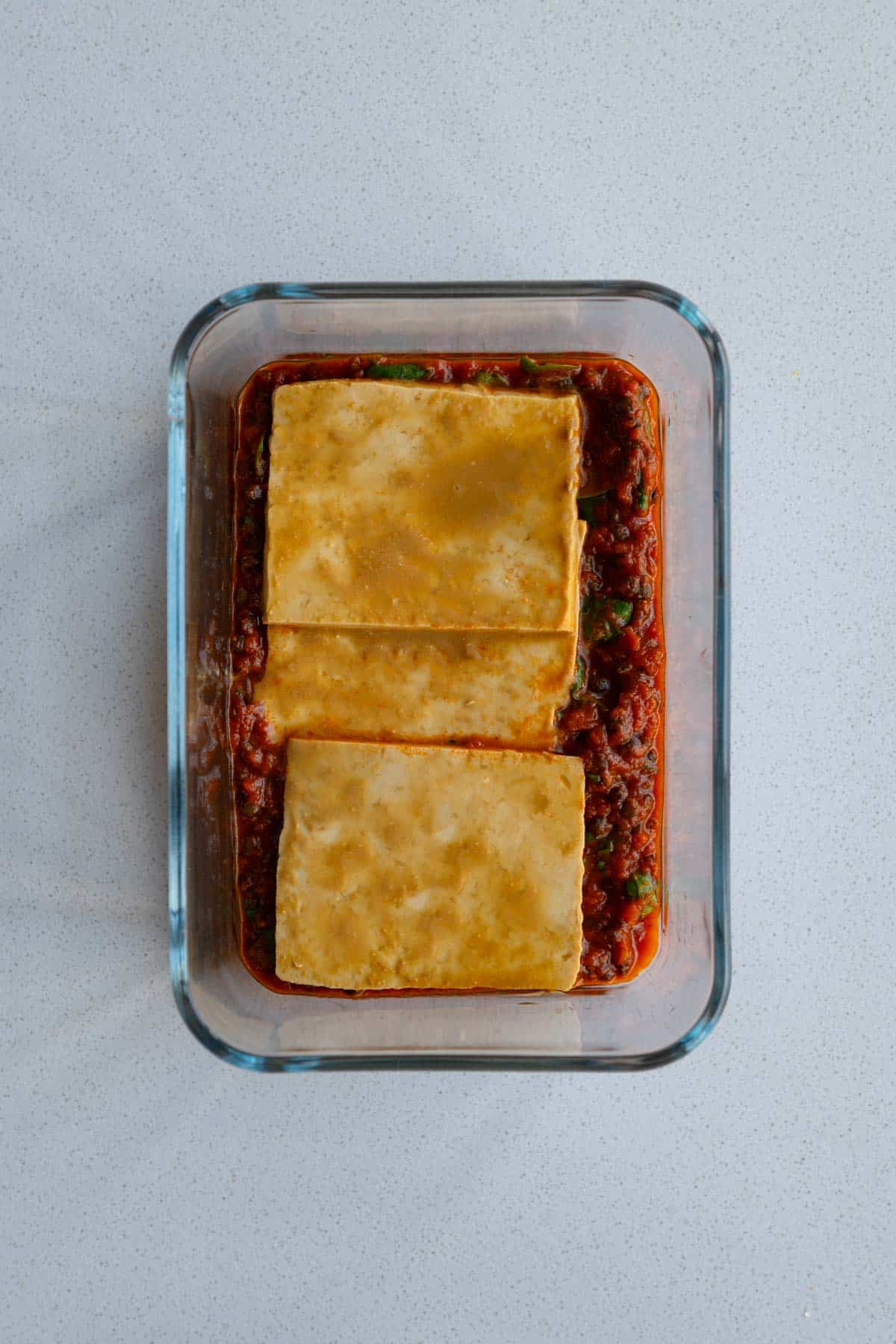 Marinara sauce with lentils and spinach with sliced marinating tofu on top at the bottom of a baking dish.