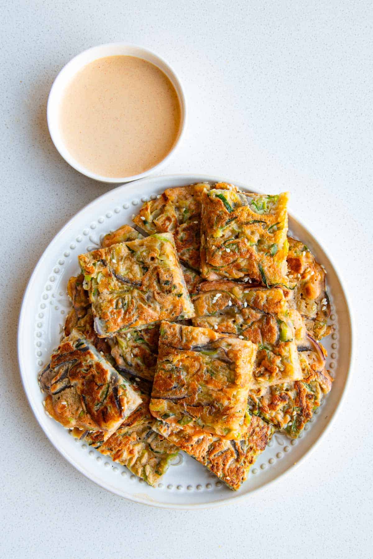 Zucchini pancakes chopped into squares on a white plate with a dipping sauce.