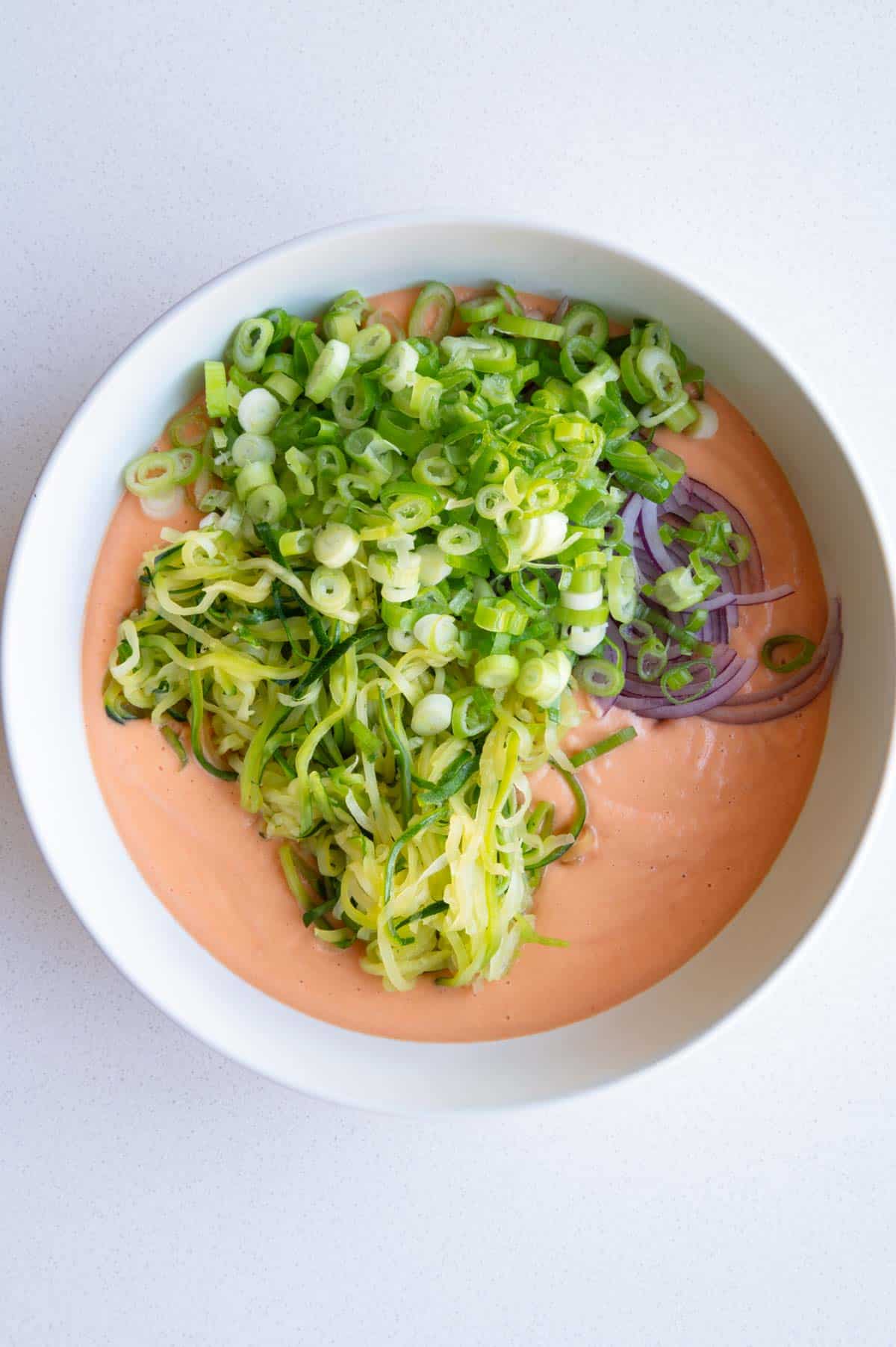 Red lentil batter in a bowl with chopped scallions, julienned zucchini, and sliced red onion.