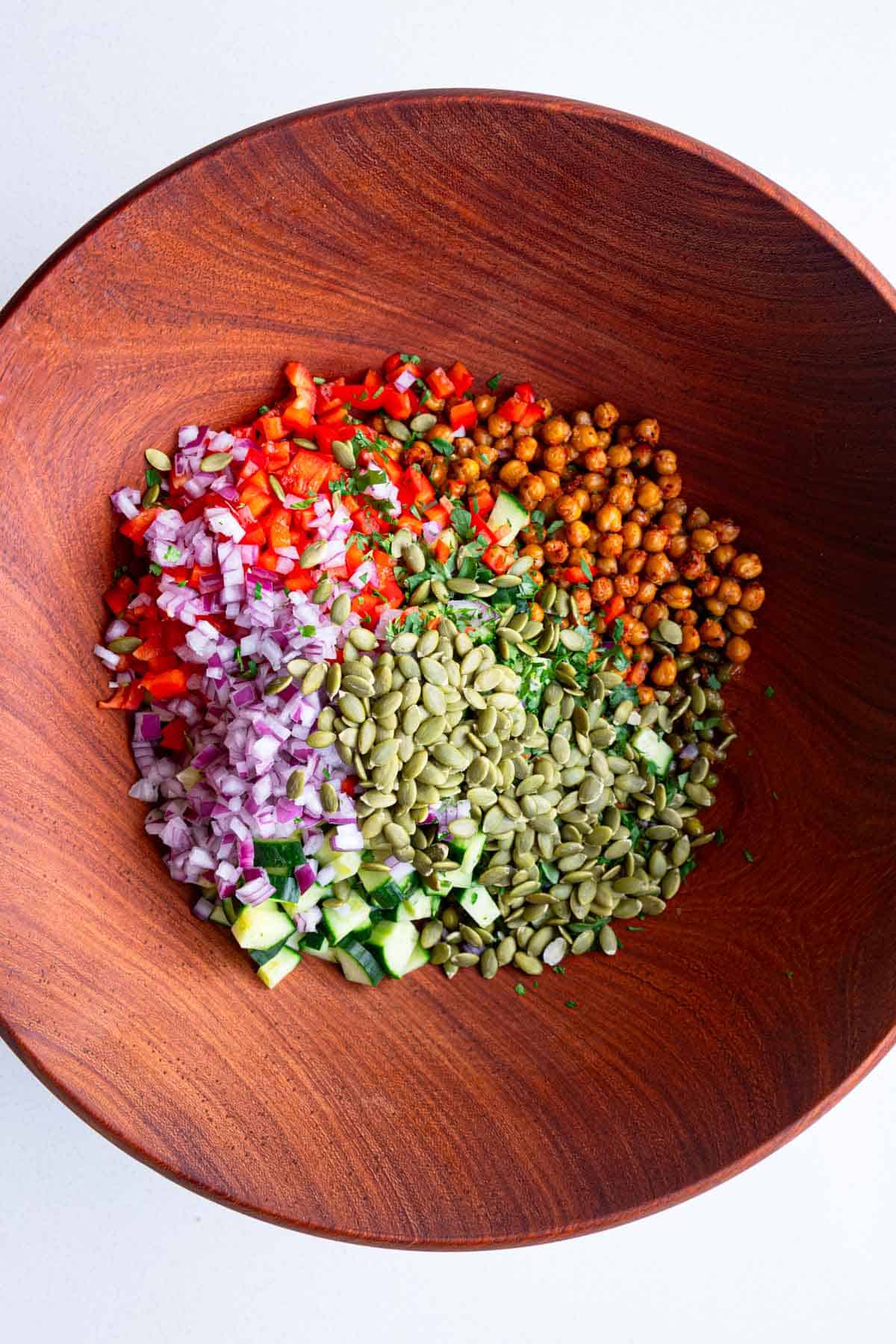 Roasted chickpeas and edamame, chopped red onion, chopped red bell pepper, pumpkin seeds, chopped cucumber, and cilantro in a wooden bowl.