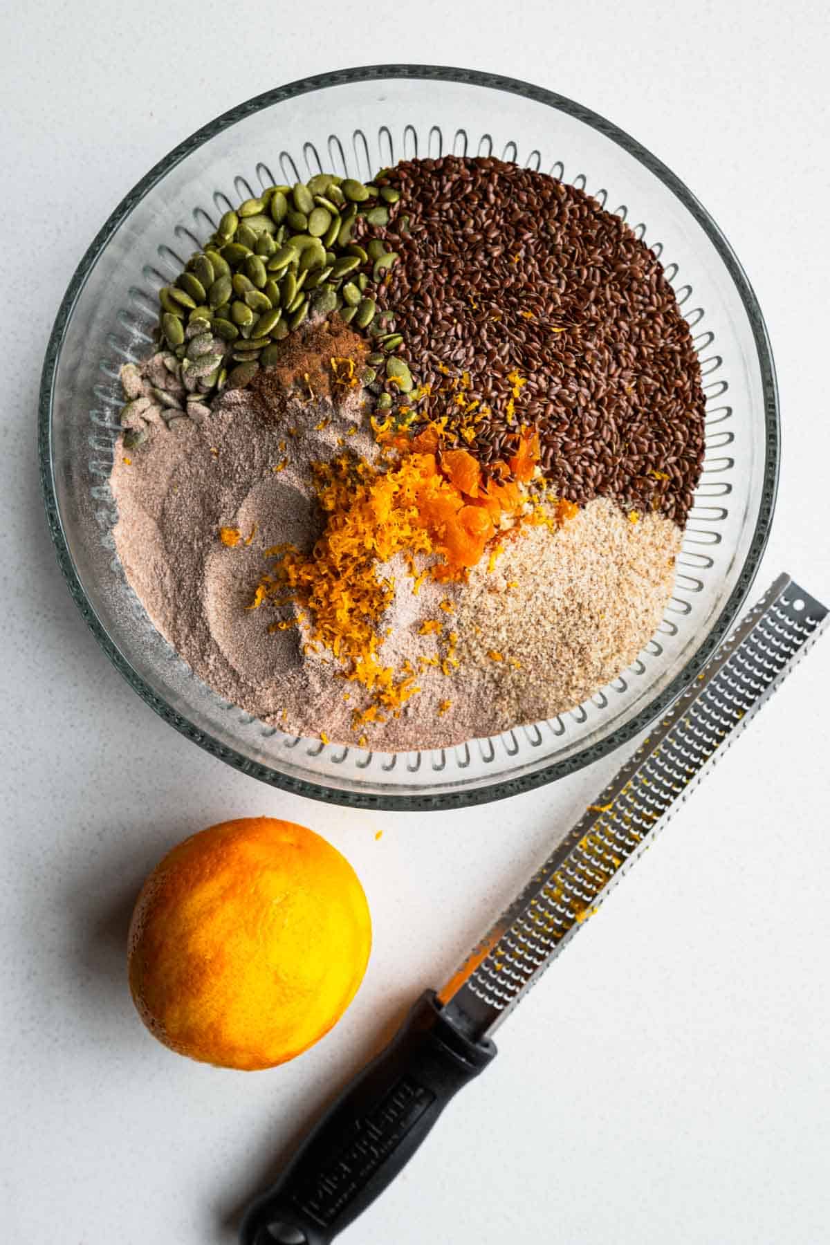 Chopped dried apricot, soaked steel-cut oats, pumpkin seeds, flaxseeds, teff flour, psyllium husk, cinnamon, and orange zest in a glass bowl.