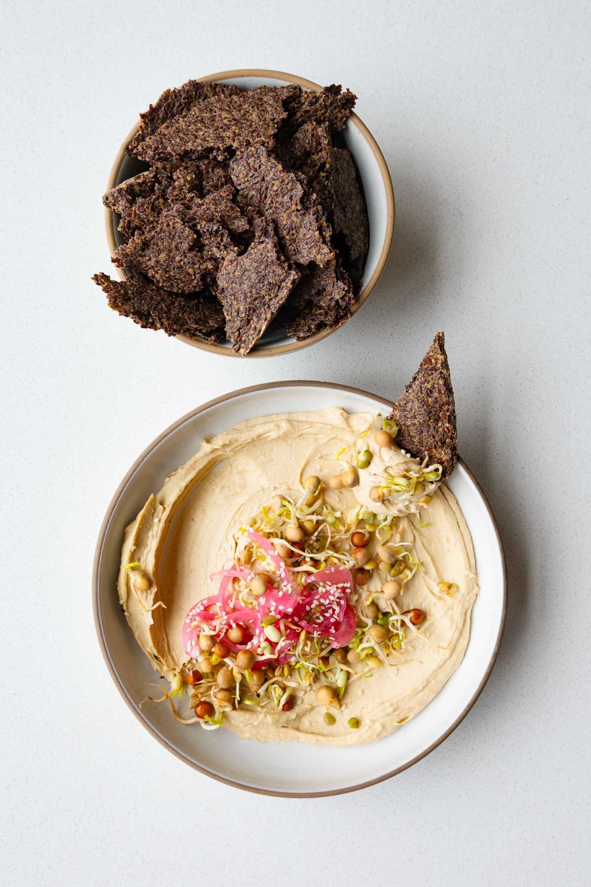 Ground flaxseed crackers in a bowl with a side of hummus and sprouts.