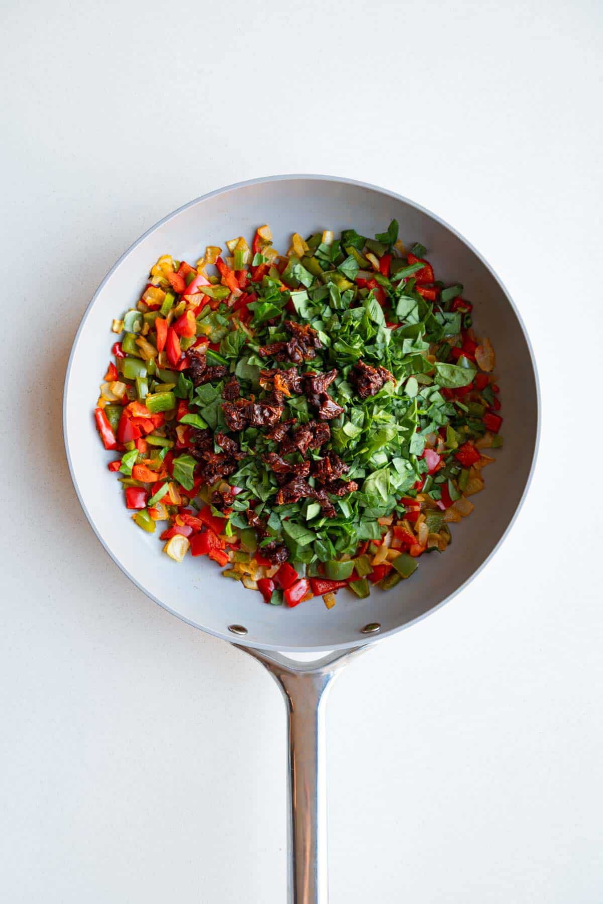Cooked chopped red and green bell peppers and onion with chopped spinach and sun-dried tomatoes in a saucepan.