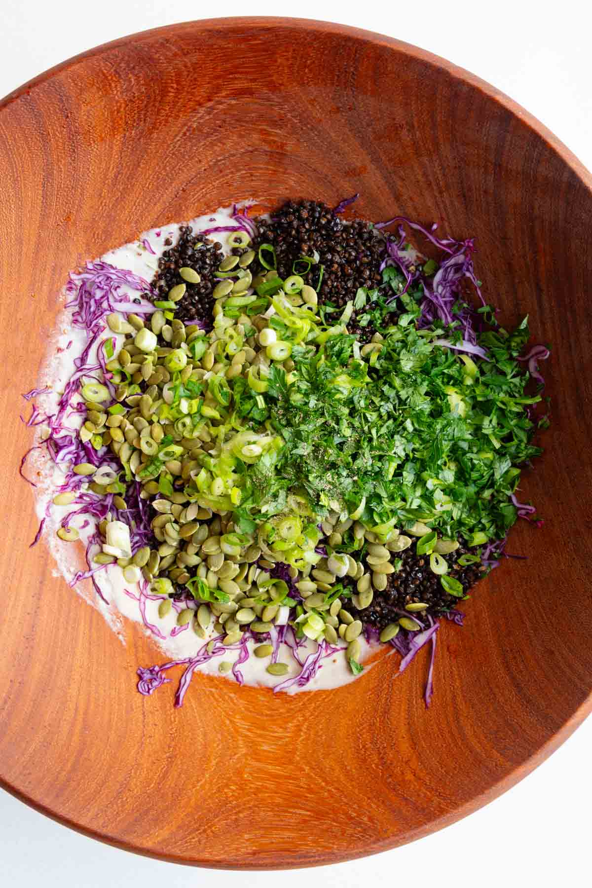 Tahini dressing, red cabbage, roasted black lentils, parsley, scallions, pumpkin seeds, and black pepper in a wooden bowl.