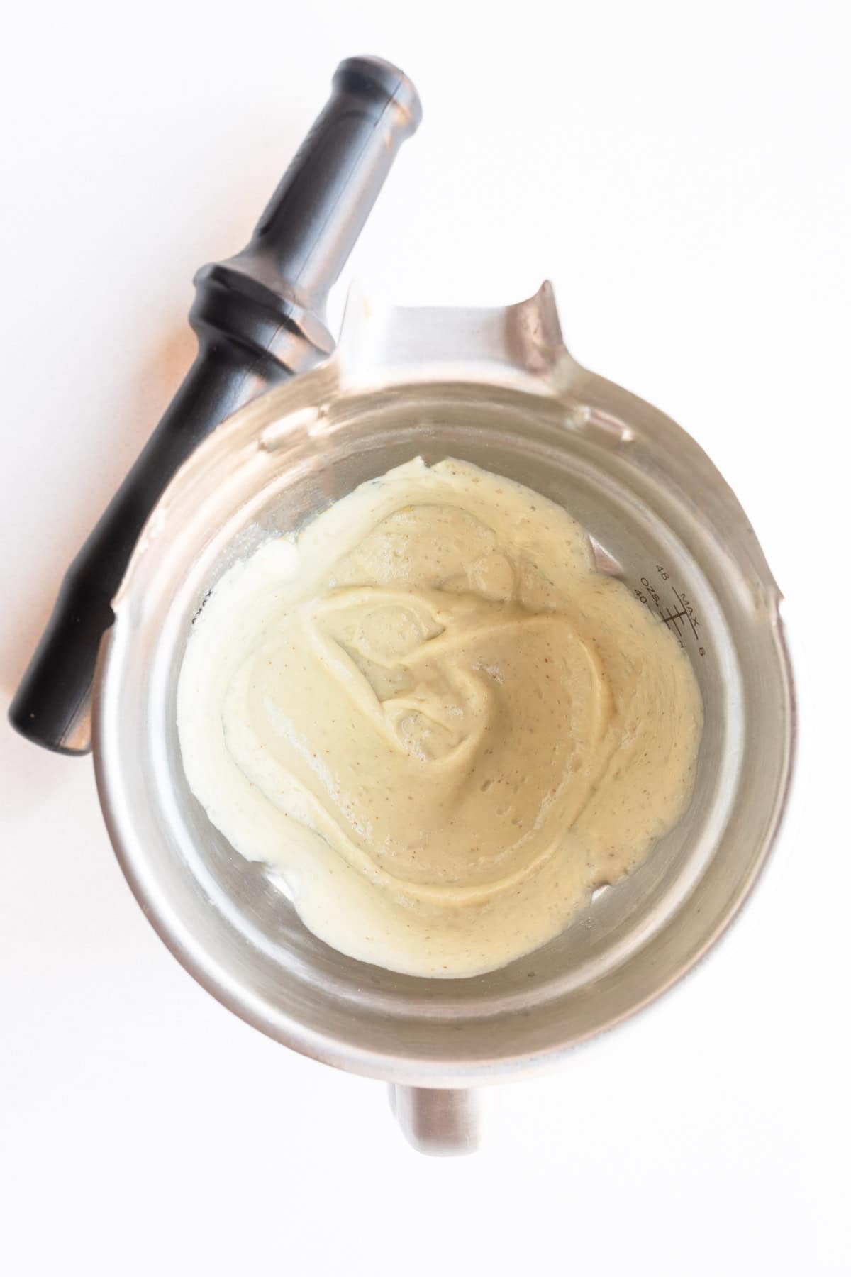 Blended white sauce in a blender next to a tamper.