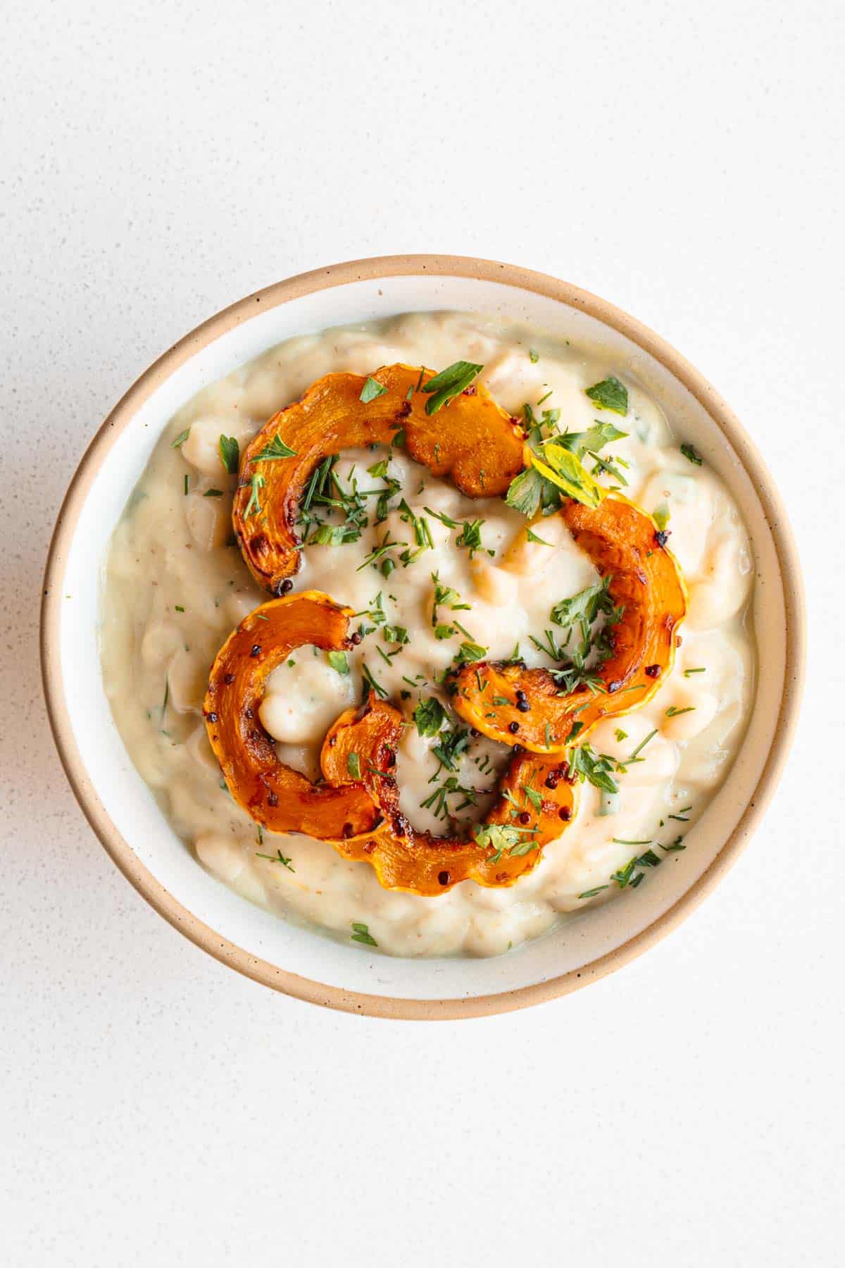 White beans in cauliflower cream sauce in a bowl topped with Delicata squash and fresh herbs.